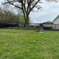 Lawn with black fence, swing set, and a large tree in the background under an overcast sky.
