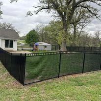 Black metal fence surrounding a yard with a house, grass, and trees.