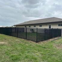 Black metal fence surrounds a grassy yard, with a light-colored house in the background under a cloudy sky.