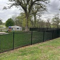 Black metal fence surrounding a grassy yard with a small building and large tree in the background.