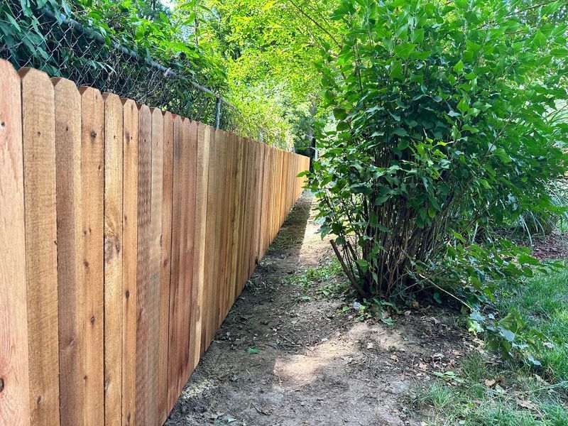 Wooden fence with decorative top next to a green bush and chain-link fence.