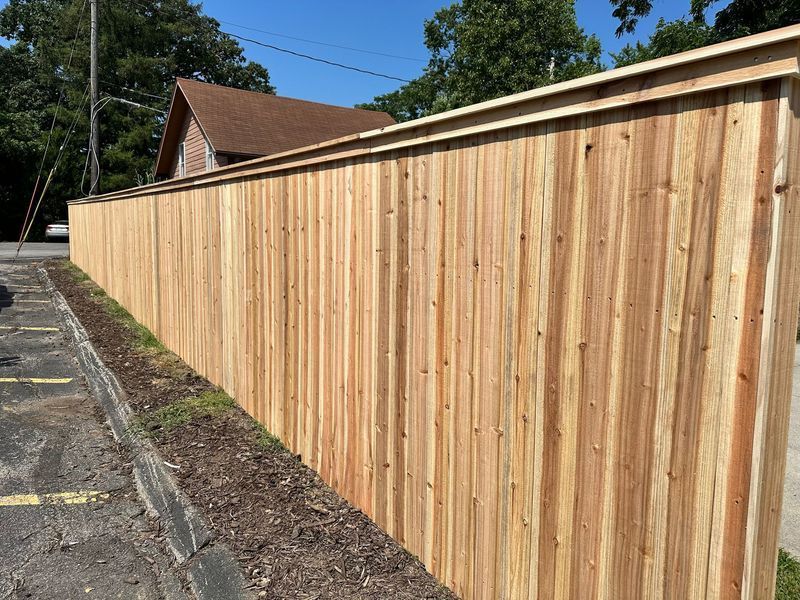 Wooden privacy fence alongside a parking lot, with a building's roof visible in the background.