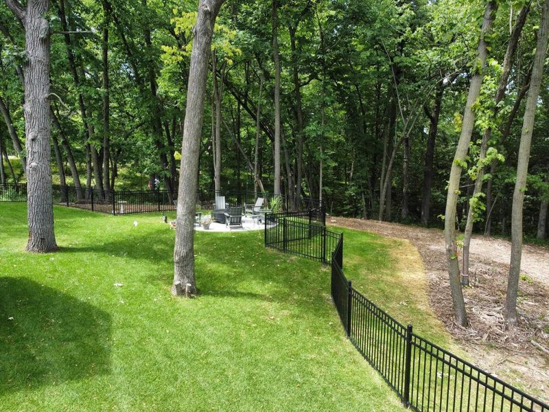 Green lawn slopes down to a black fence, with trees and a patio in the background.