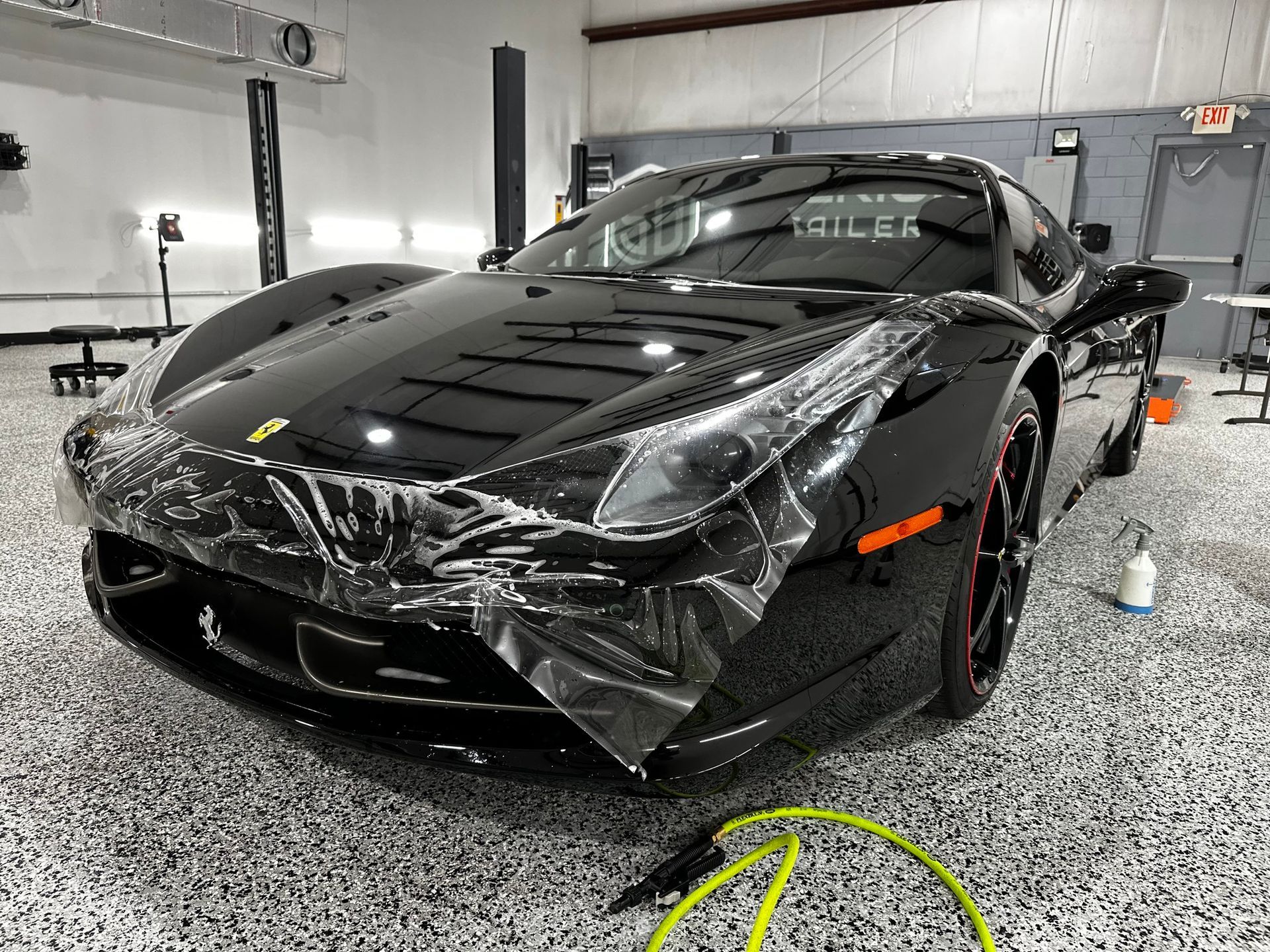 A black sports car is wrapped in plastic in a garage.