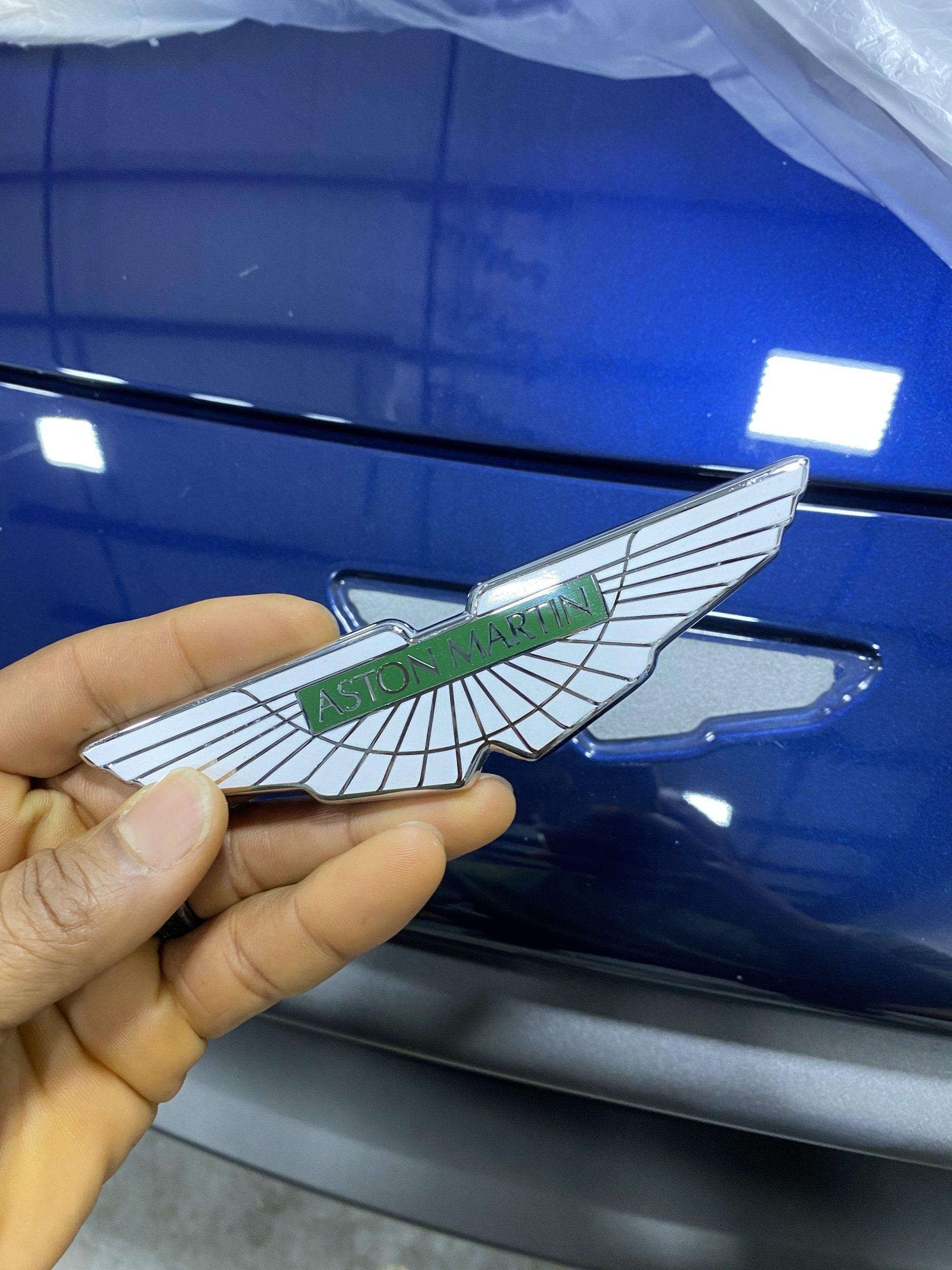 A person is holding a aston martin emblem in front of a blue car