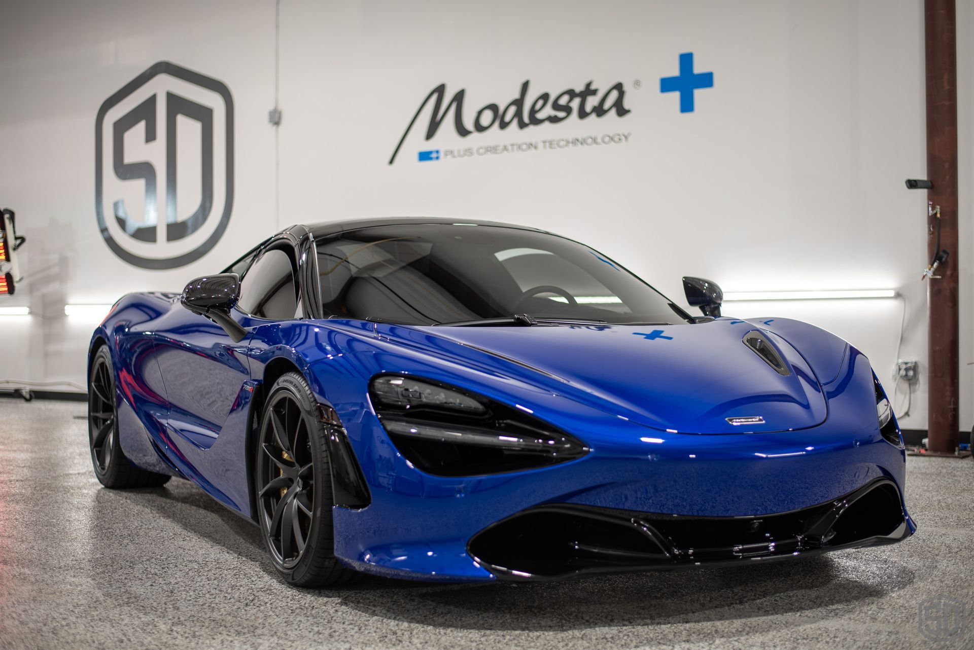 A blue mclaren 720s is parked in a garage in front of a white wall.