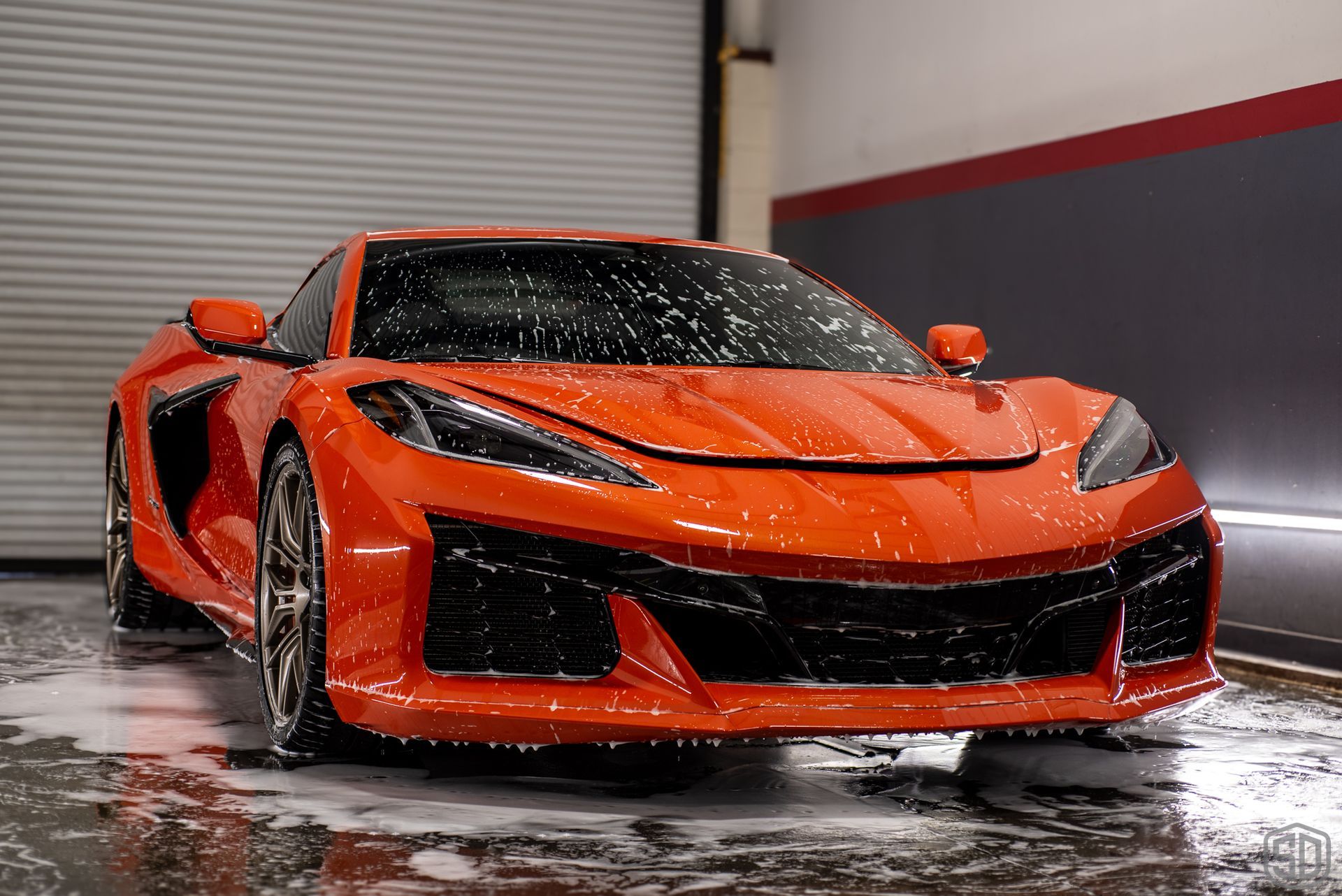 Orange sports car being washed, covered in soap suds, in a garage setting.