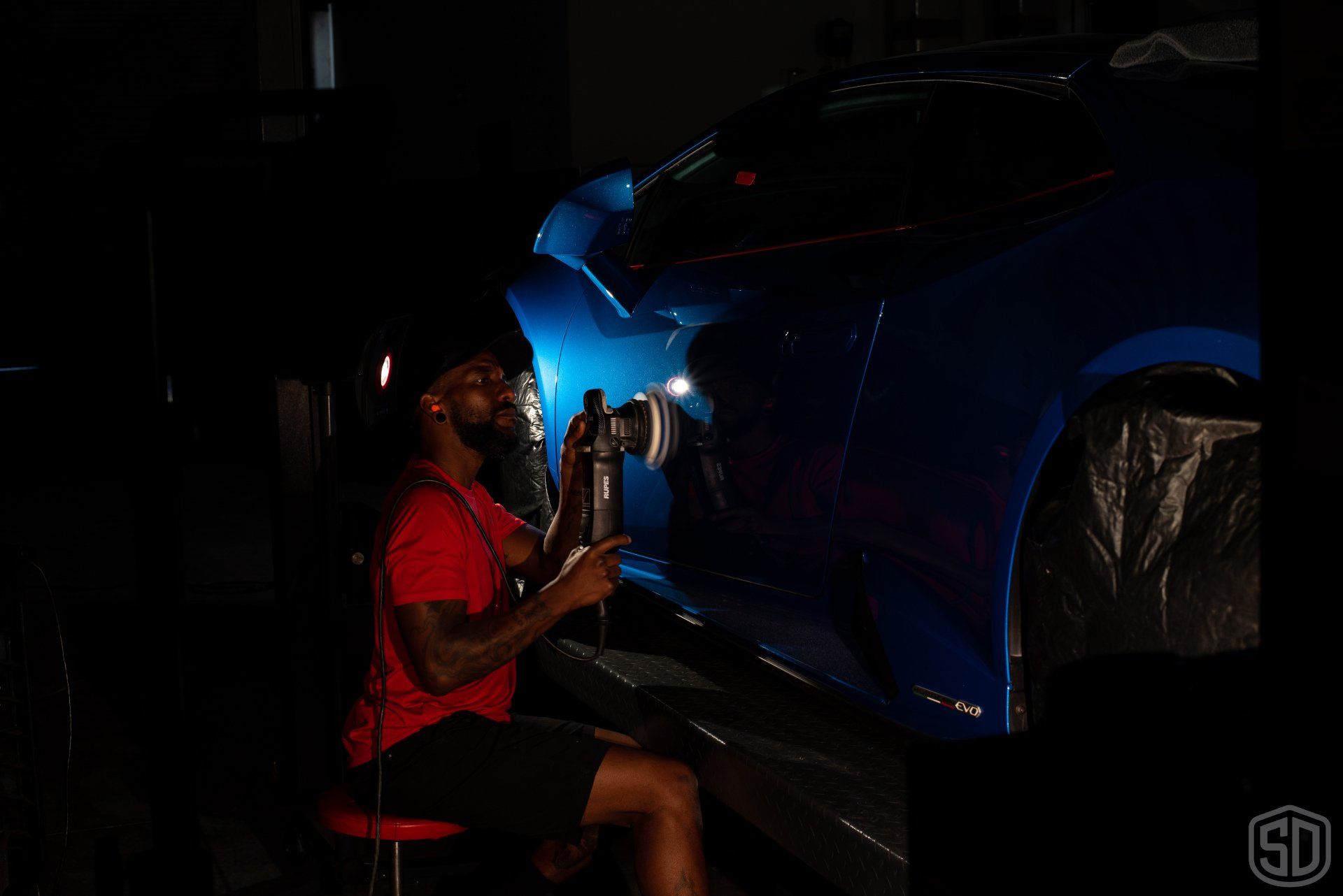 A man is sitting on a stool in front of a blue car in a dark room.