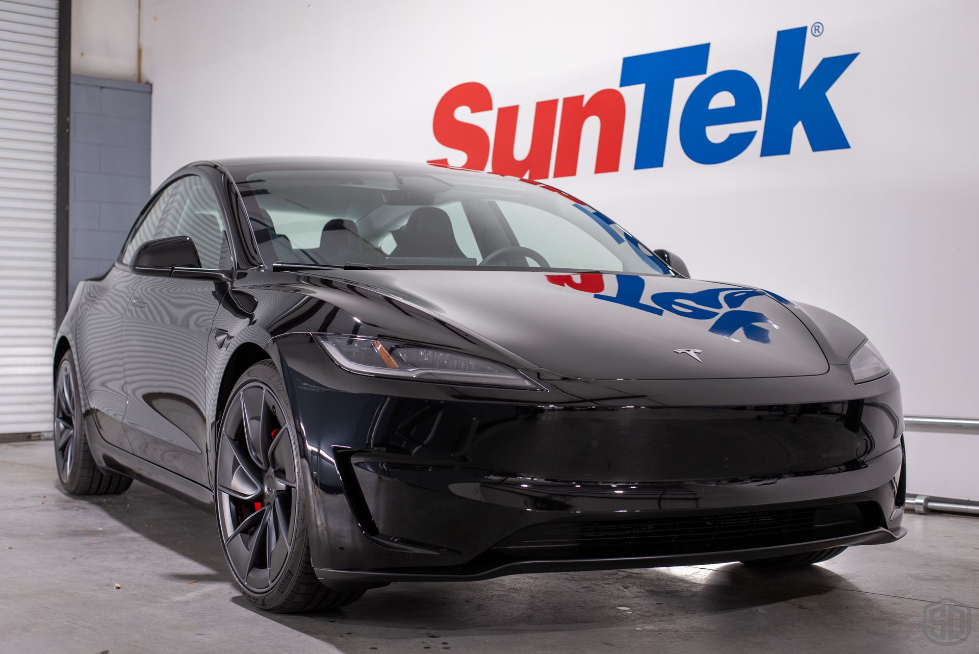 A black tesla model 3 is parked in front of a suntek sign.