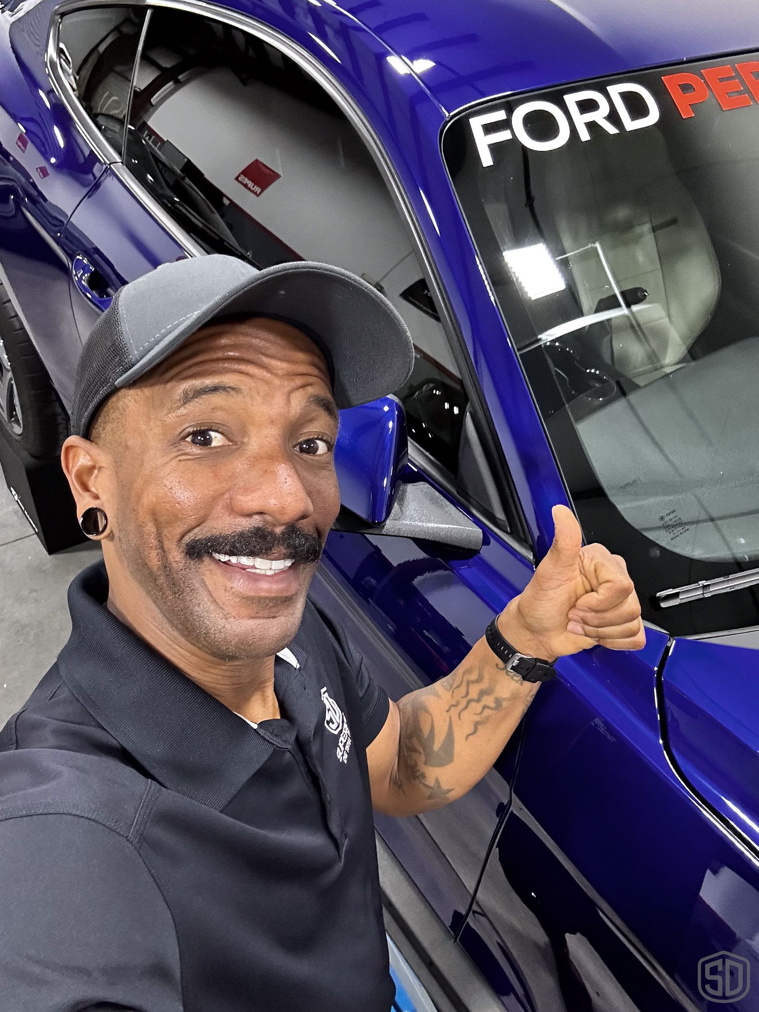 A man is giving a thumbs up in front of a blue ford mustang.