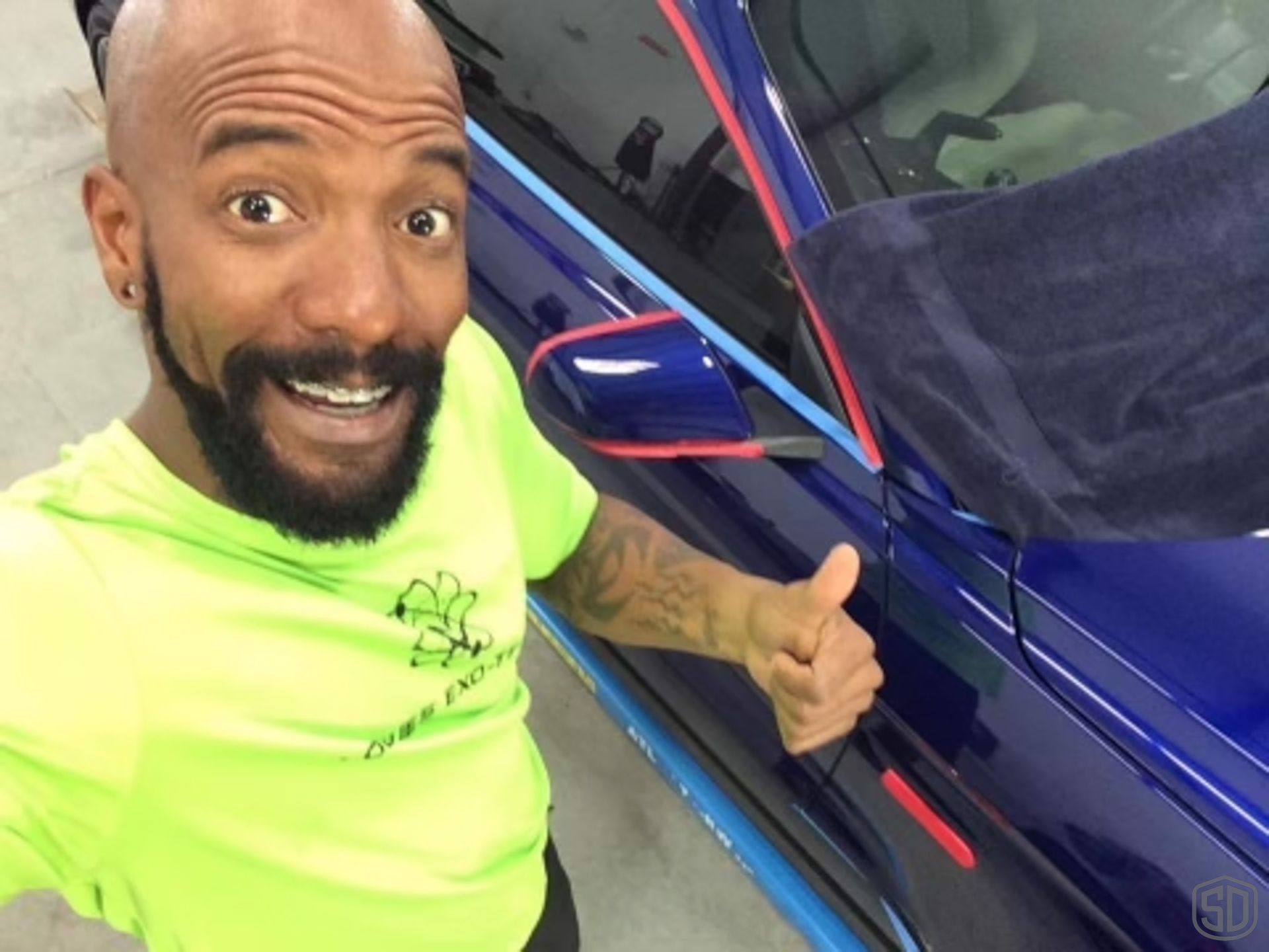 A man with a beard is giving a thumbs up in front of a blue car