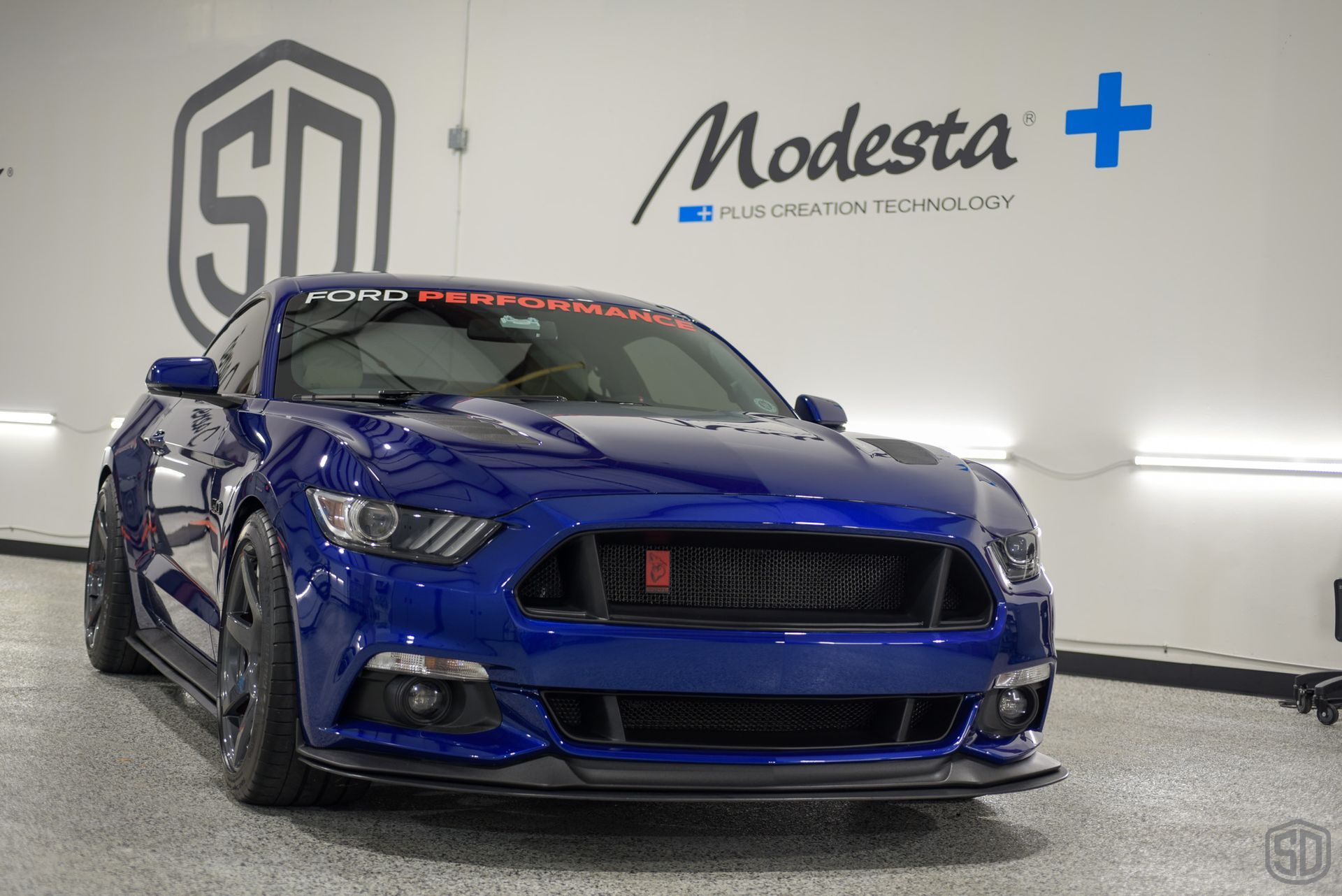 A blue mustang is parked in a garage in front of a wall that says modesta +.