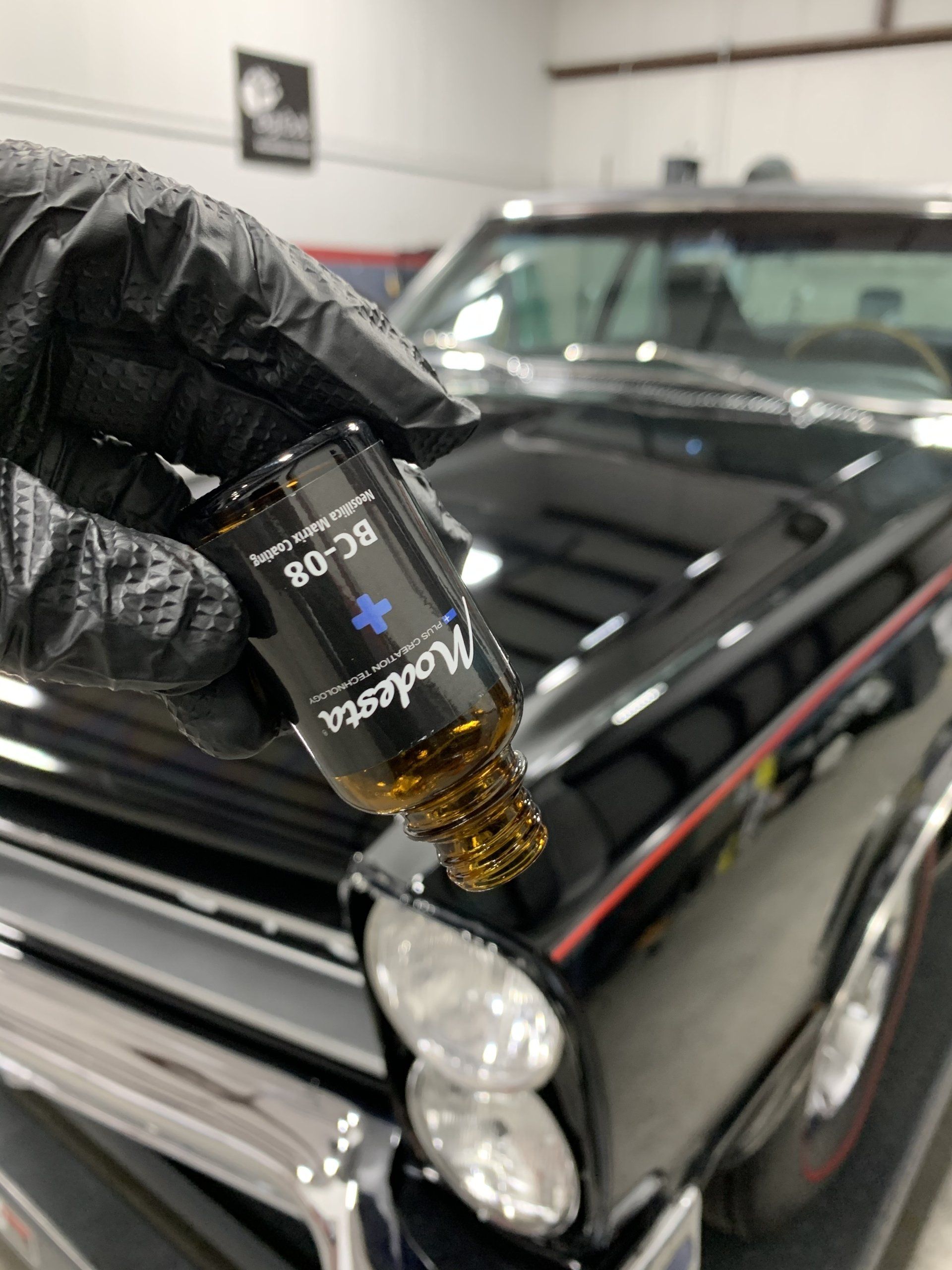 A person in black gloves is holding a bottle in front of a black car