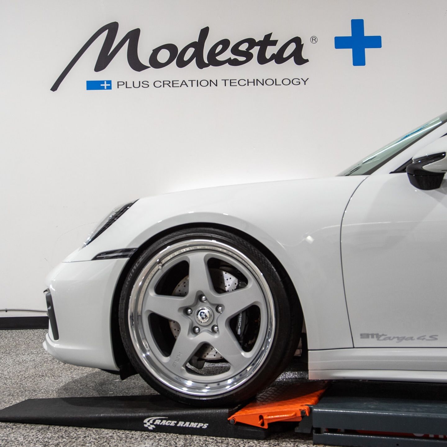 A white car is parked in front of a wall that says modesta plus creation technology