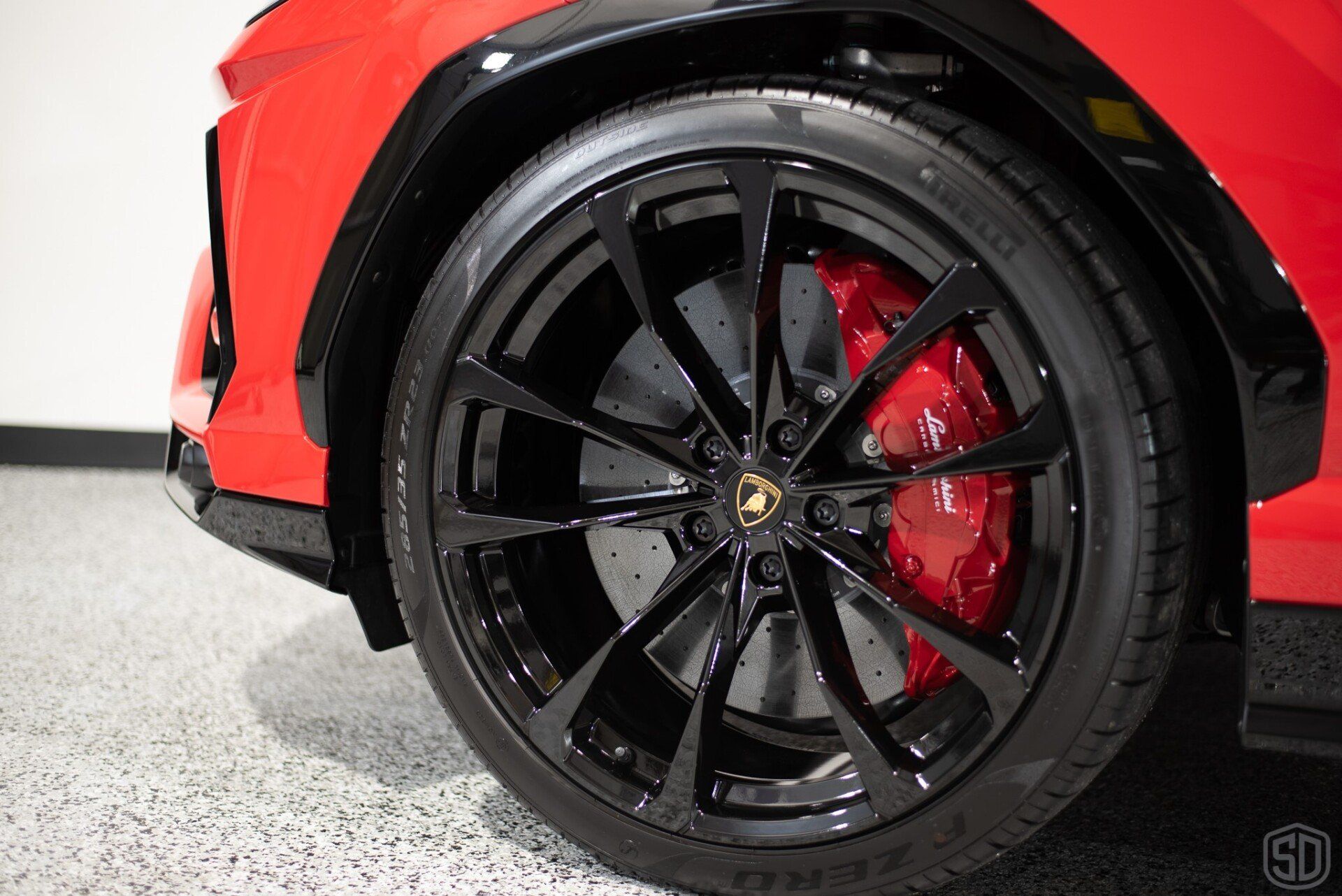 A red car with black wheels and red brake calipers is parked in a garage.
