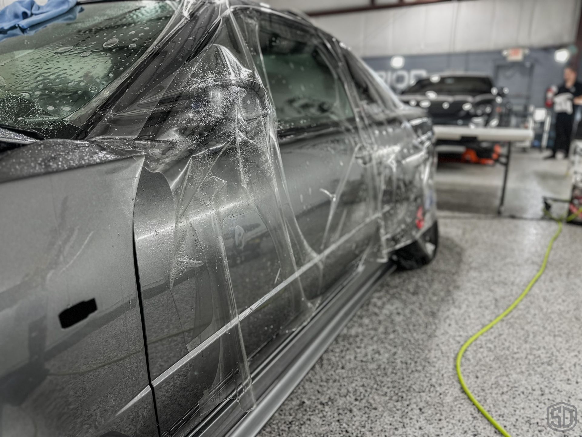 A car is being covered in plastic wrap in a garage.
