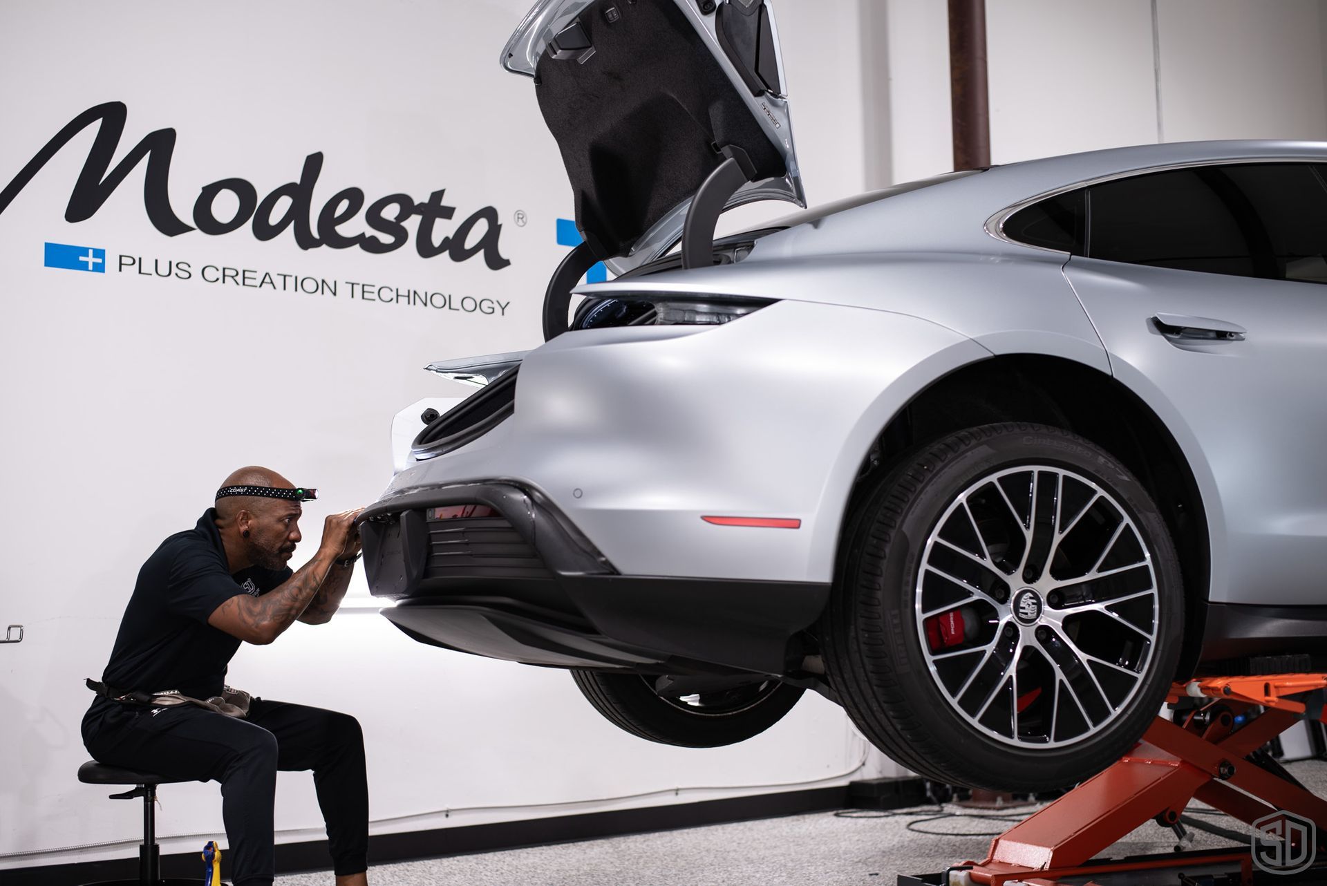 A man is sitting on a chair applying PPF to the rear bumper of a car.