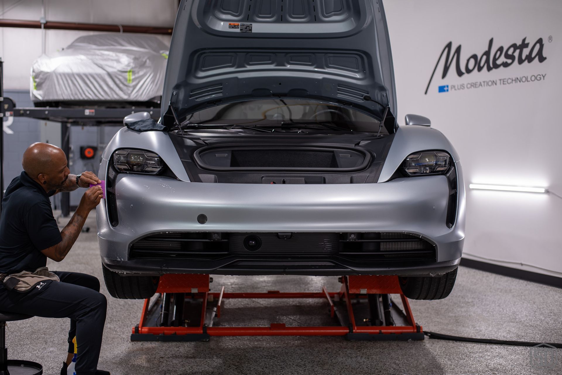 A man is sitting on a chair applying PPF to the rear bumper of a car.