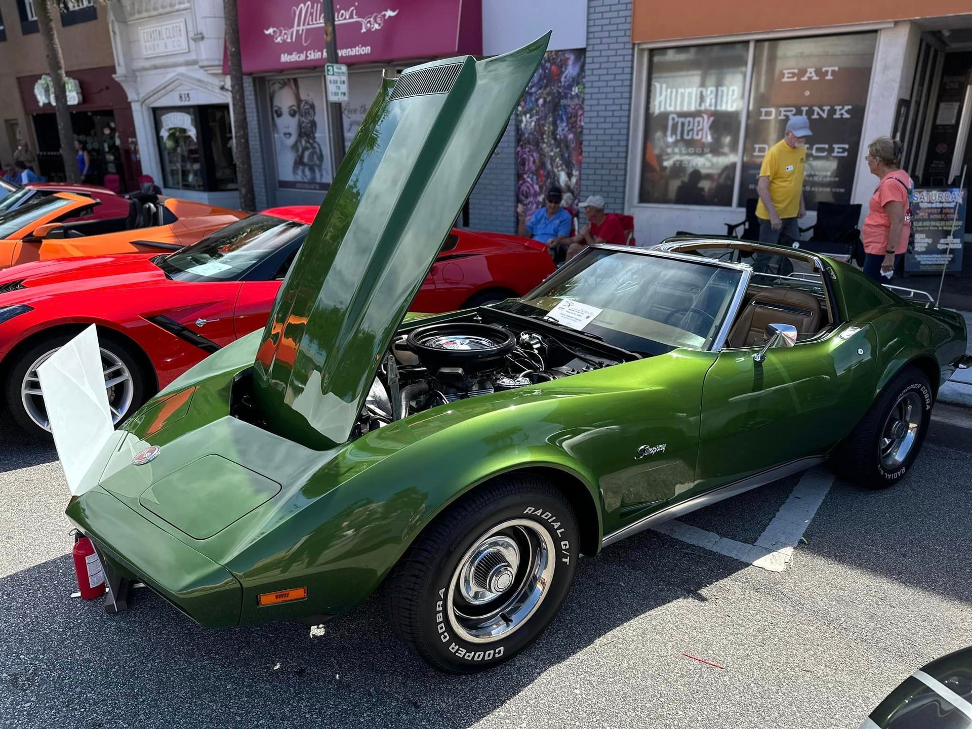 A green corvette with the hood up is parked in front of a building.