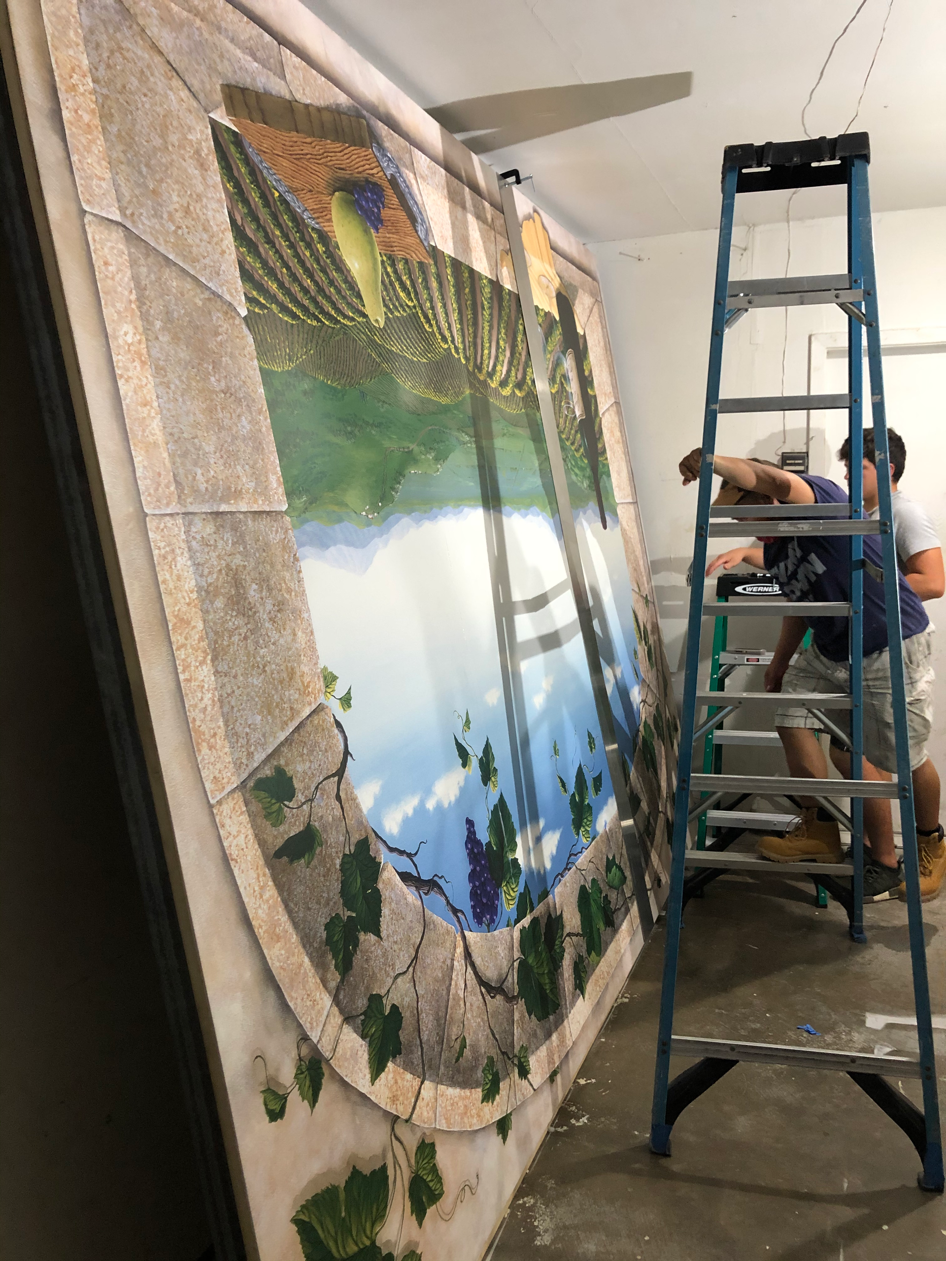 Man on ladder installing a large mural depicting a vineyard scene on a wall.