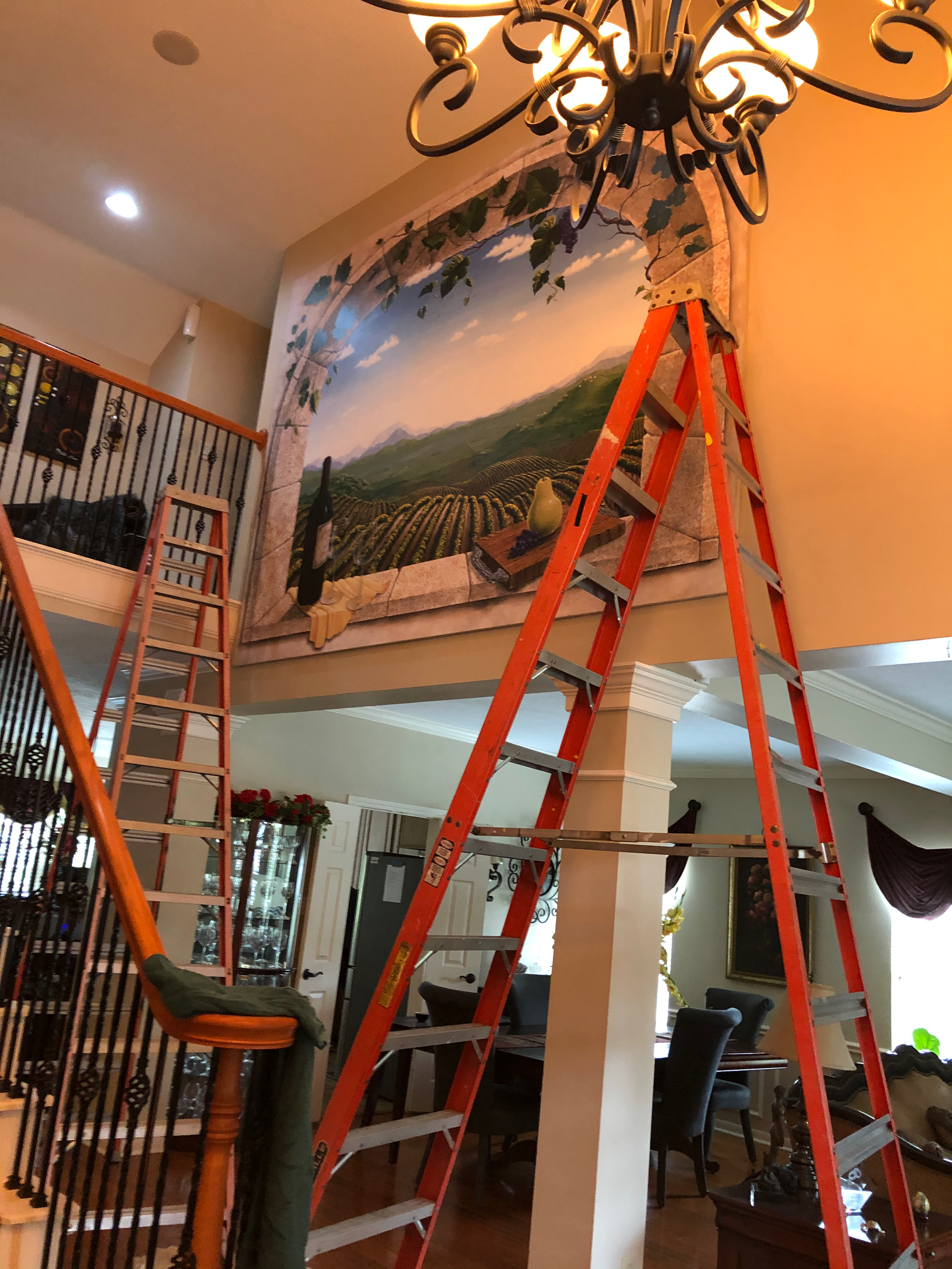A mural of a landscape is being worked on from a ladder. A staircase and chandelier are also visible.