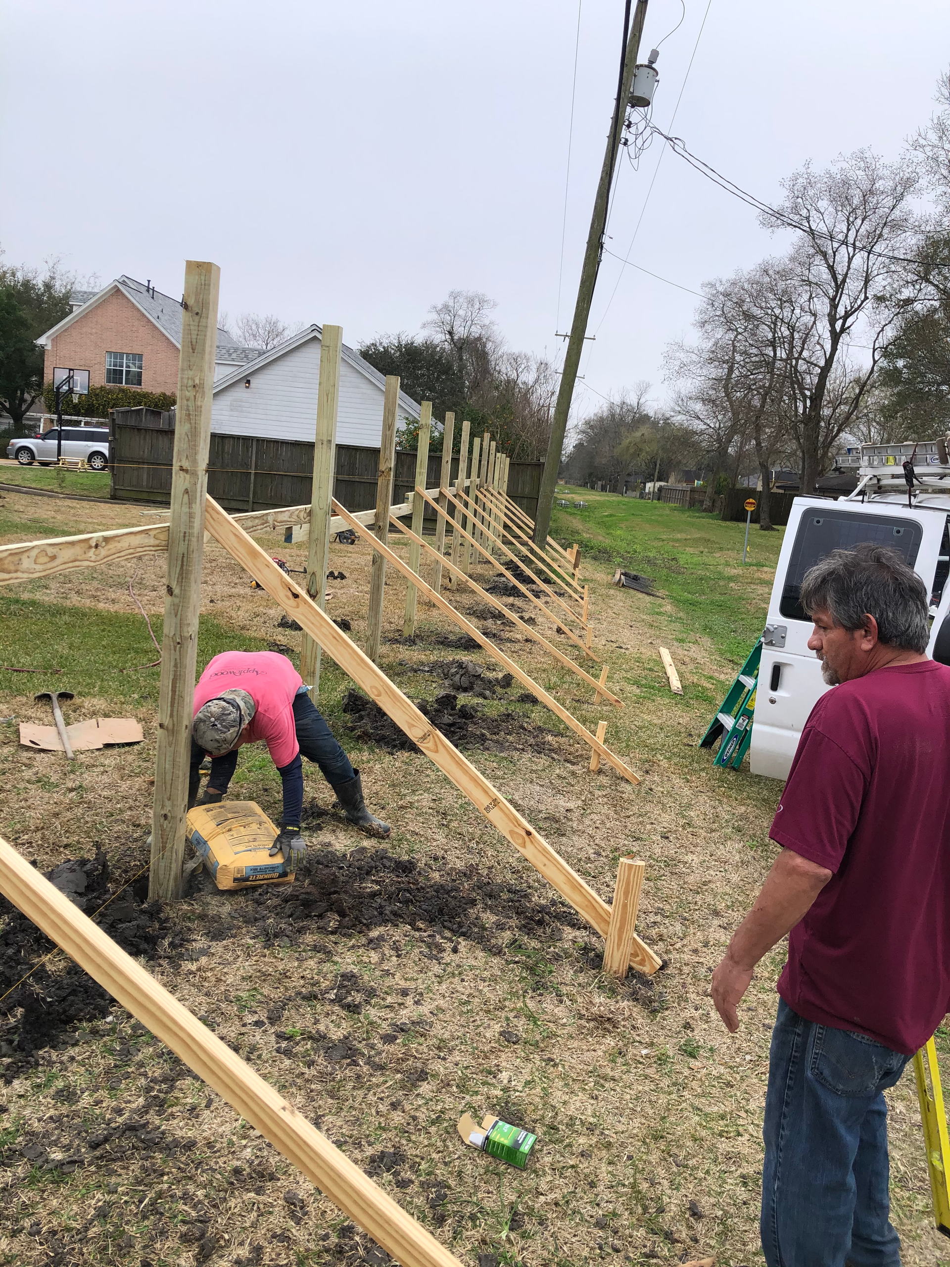 Two people building a wooden fence outdoors. One works on the ground, the other looks on.