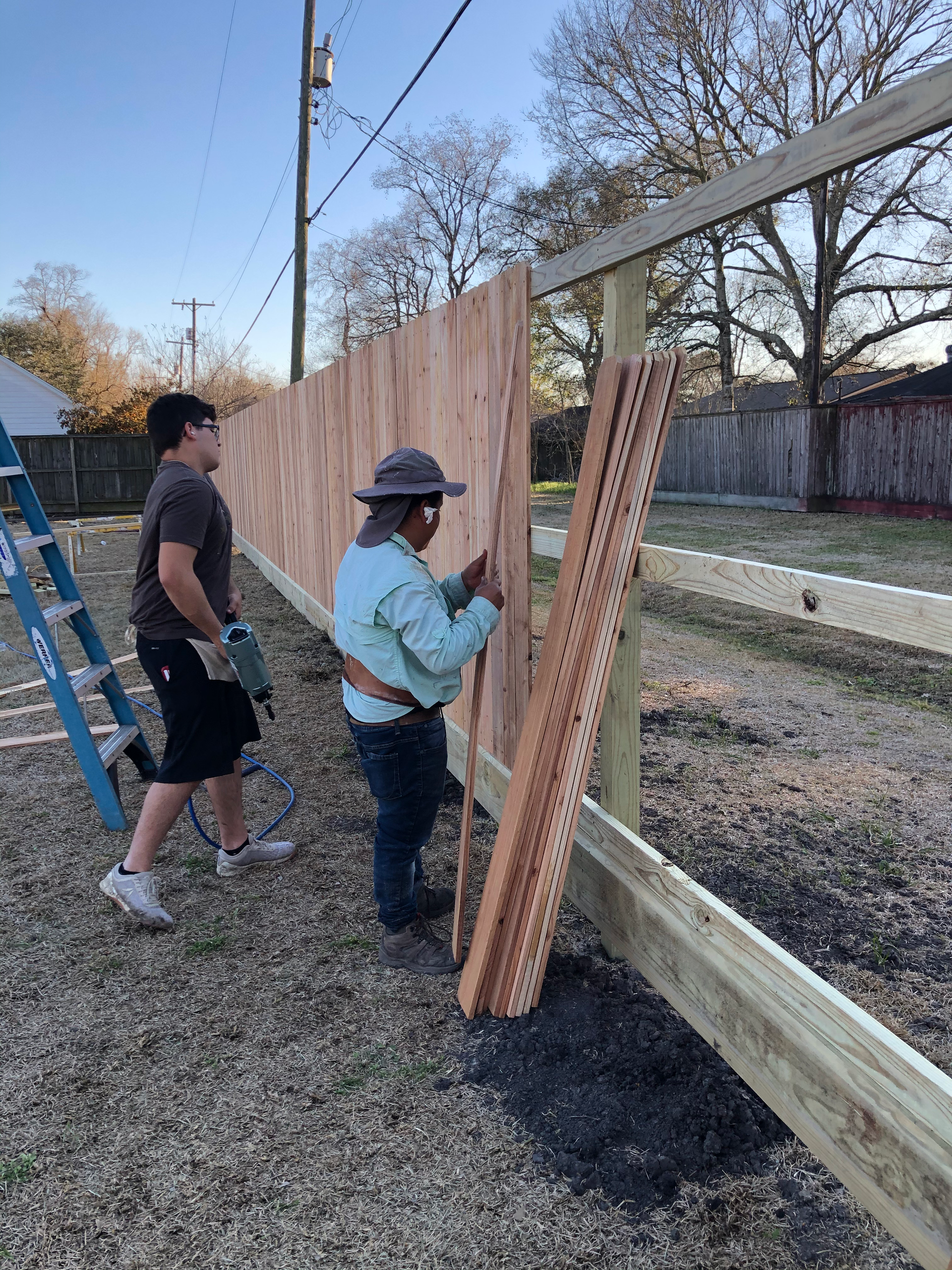 Two people building a wooden fence outdoors; one holds a nail gun, the other attaches planks.