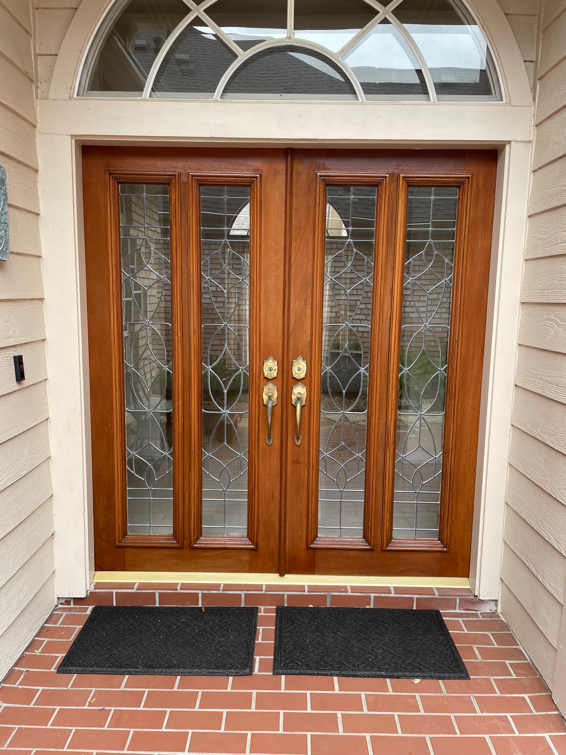 Double wooden front doors with glass panels, a brick entryway, and two black mats.