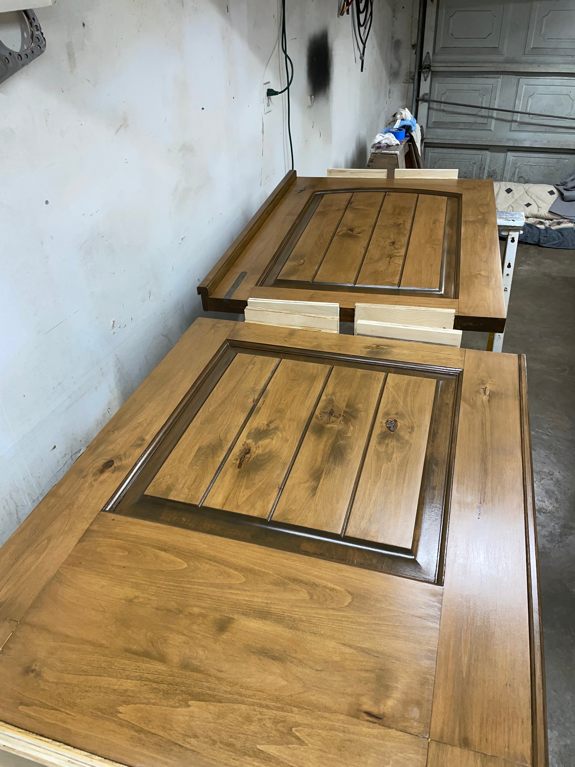 Two wooden doors, stained light brown, with dark brown accents, lying in a workshop.