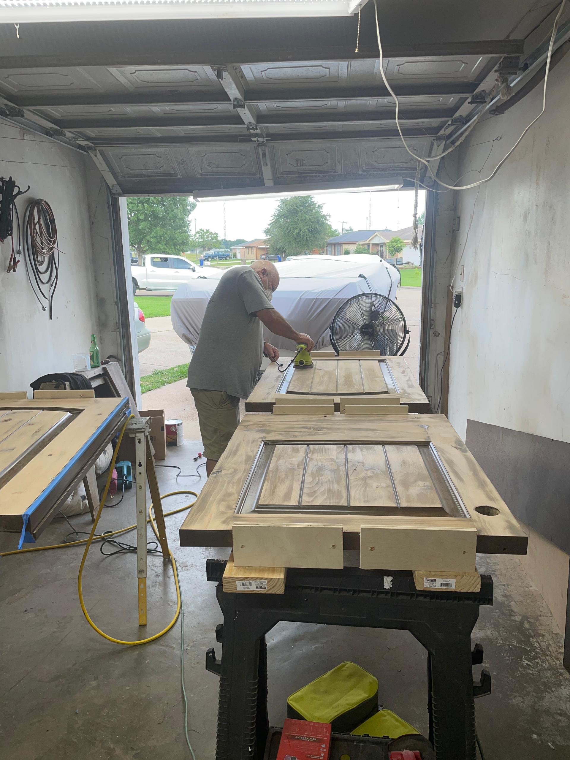 Man sanding wooden doors in a garage. Two doors are on workbenches; sunlight streams in the open garage door.