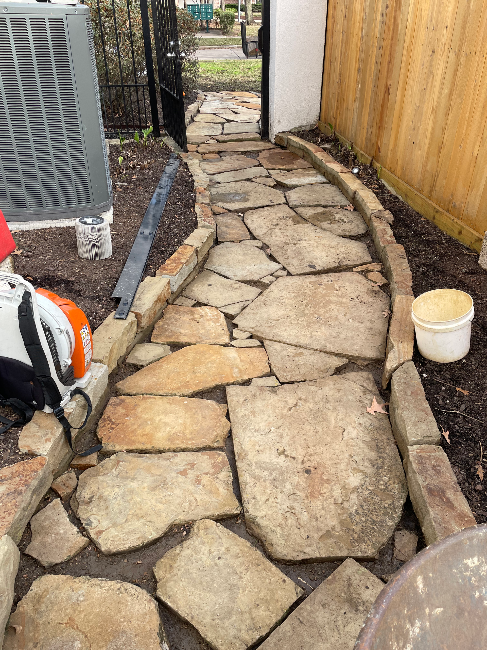 A stone pathway curves toward a gate, bordered by brick and soil. A/C unit and bucket nearby.