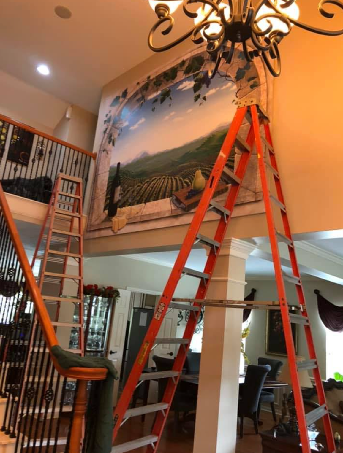 Orange ladder propped against a wall with a mural of a vineyard scene. Another ladder is near a staircase.
