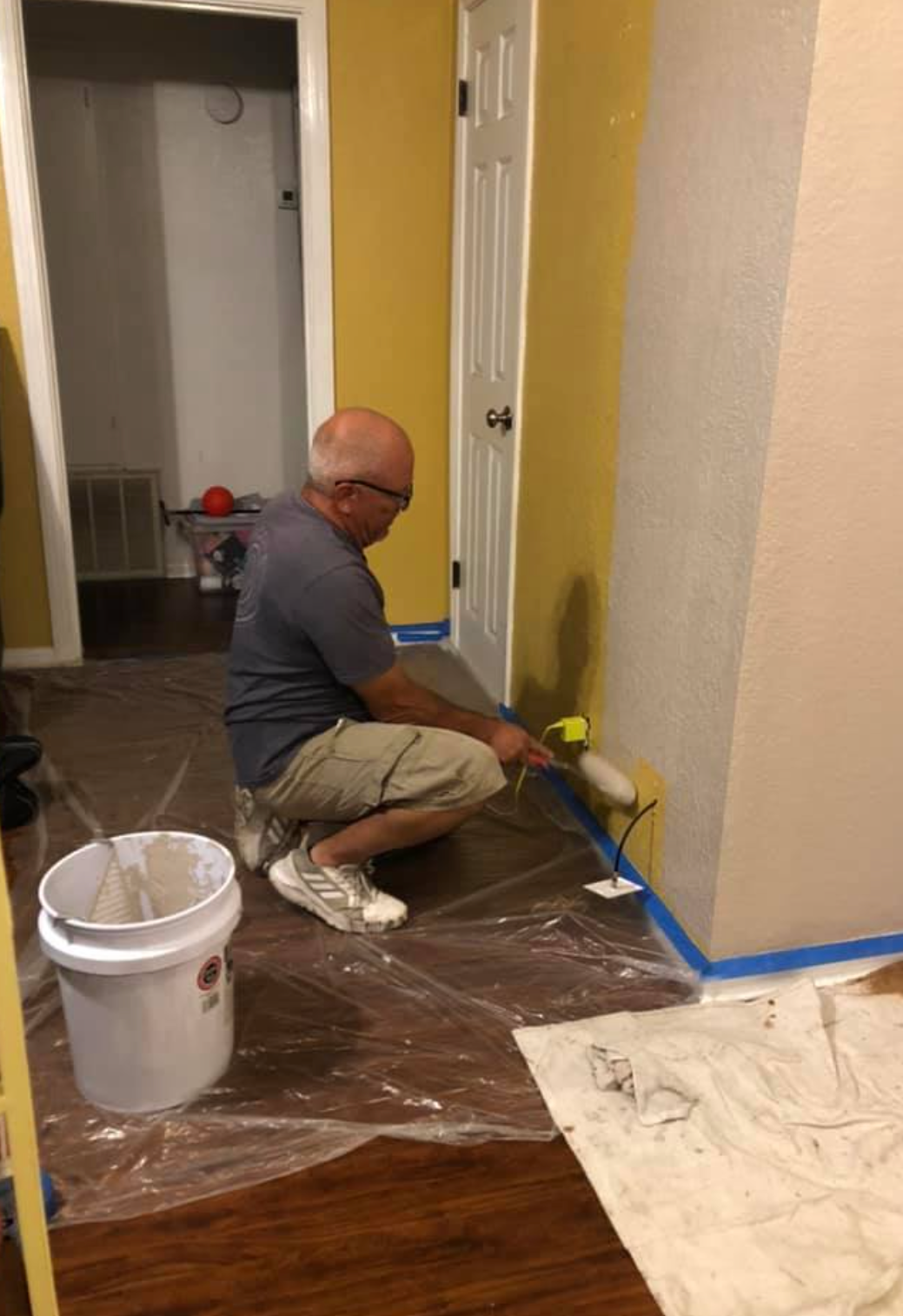 Man painting wall beige in room with yellow and white walls, brown flooring, and blue tape.