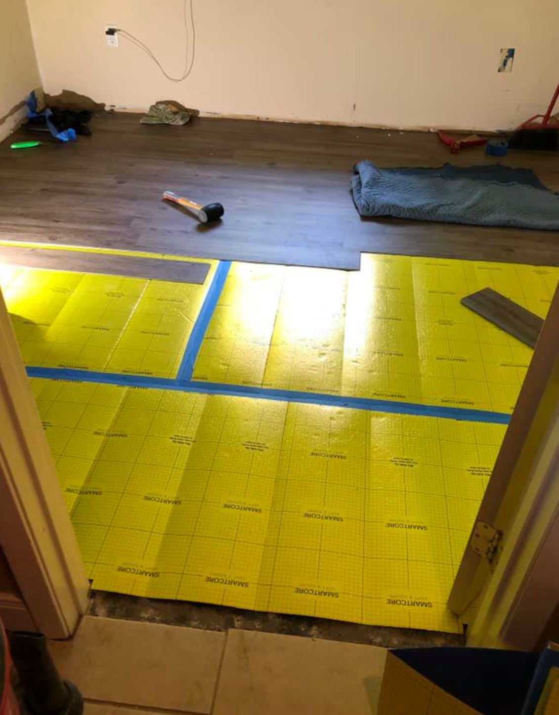 Room with yellow underlayment being installed, transitioning to existing wood flooring.