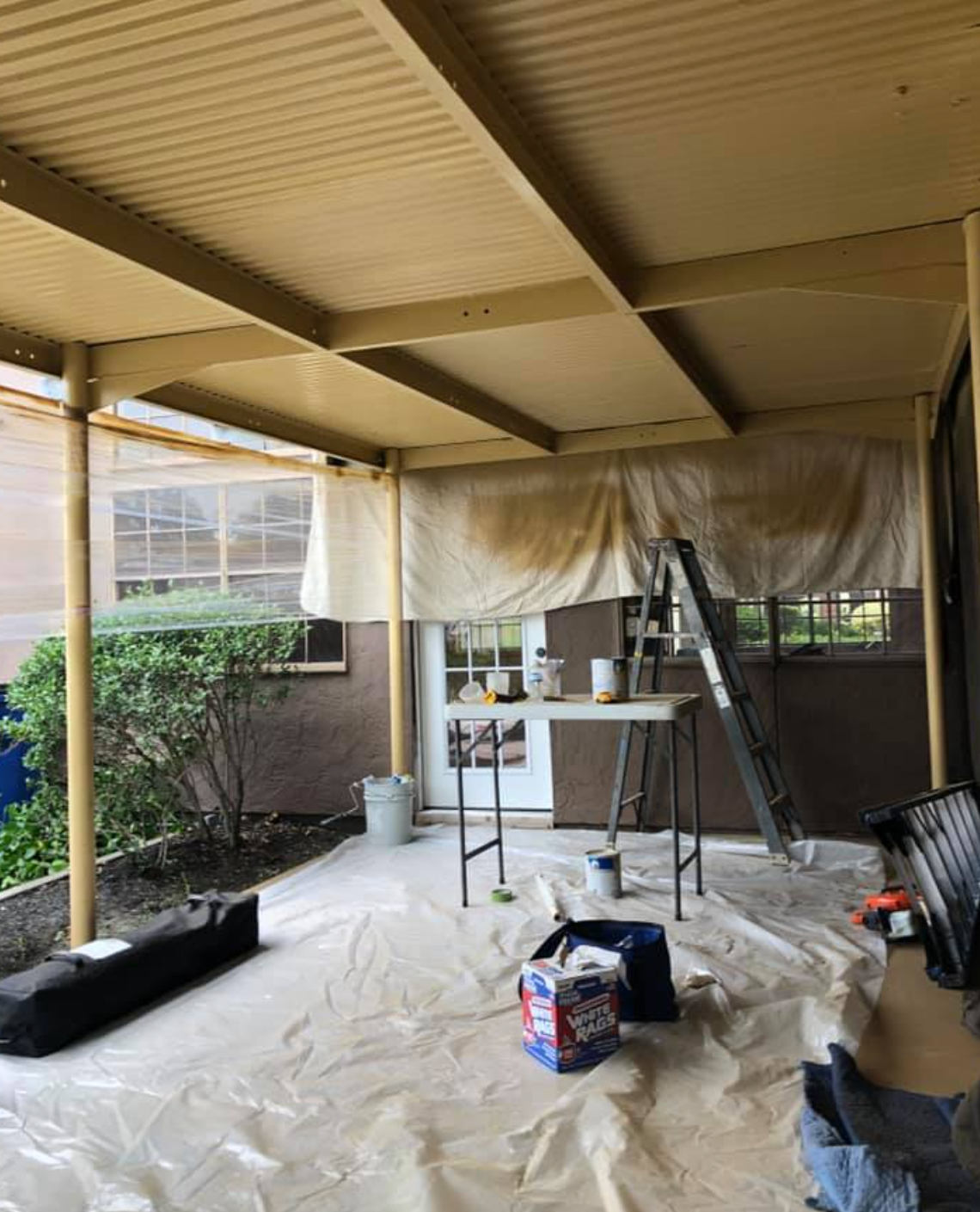 Patio under renovation with painting supplies, tarp, and ladder. Brown and tan color scheme.