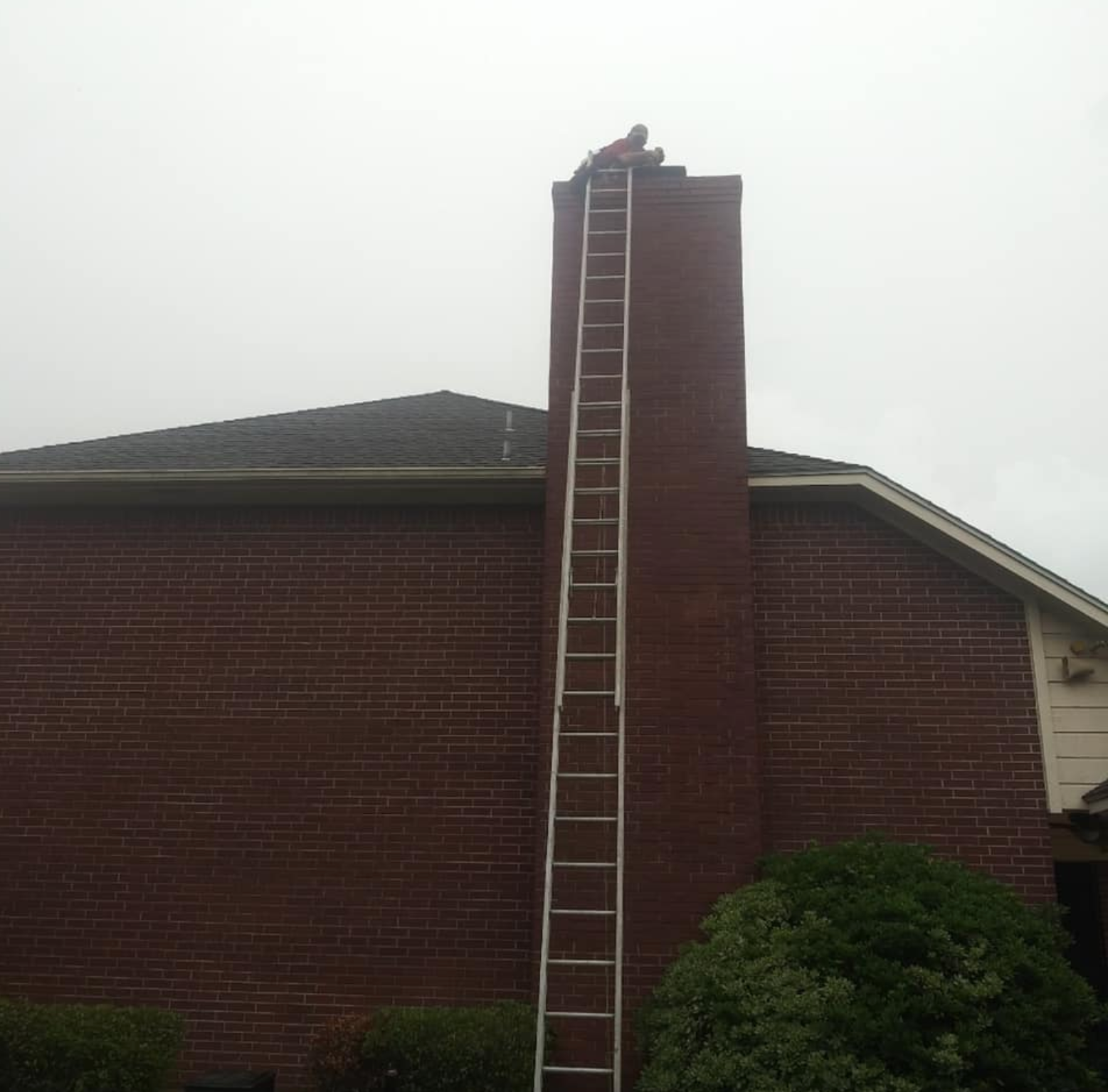 Person on a tall ladder cleaning the chimney of a brick house on a cloudy day.