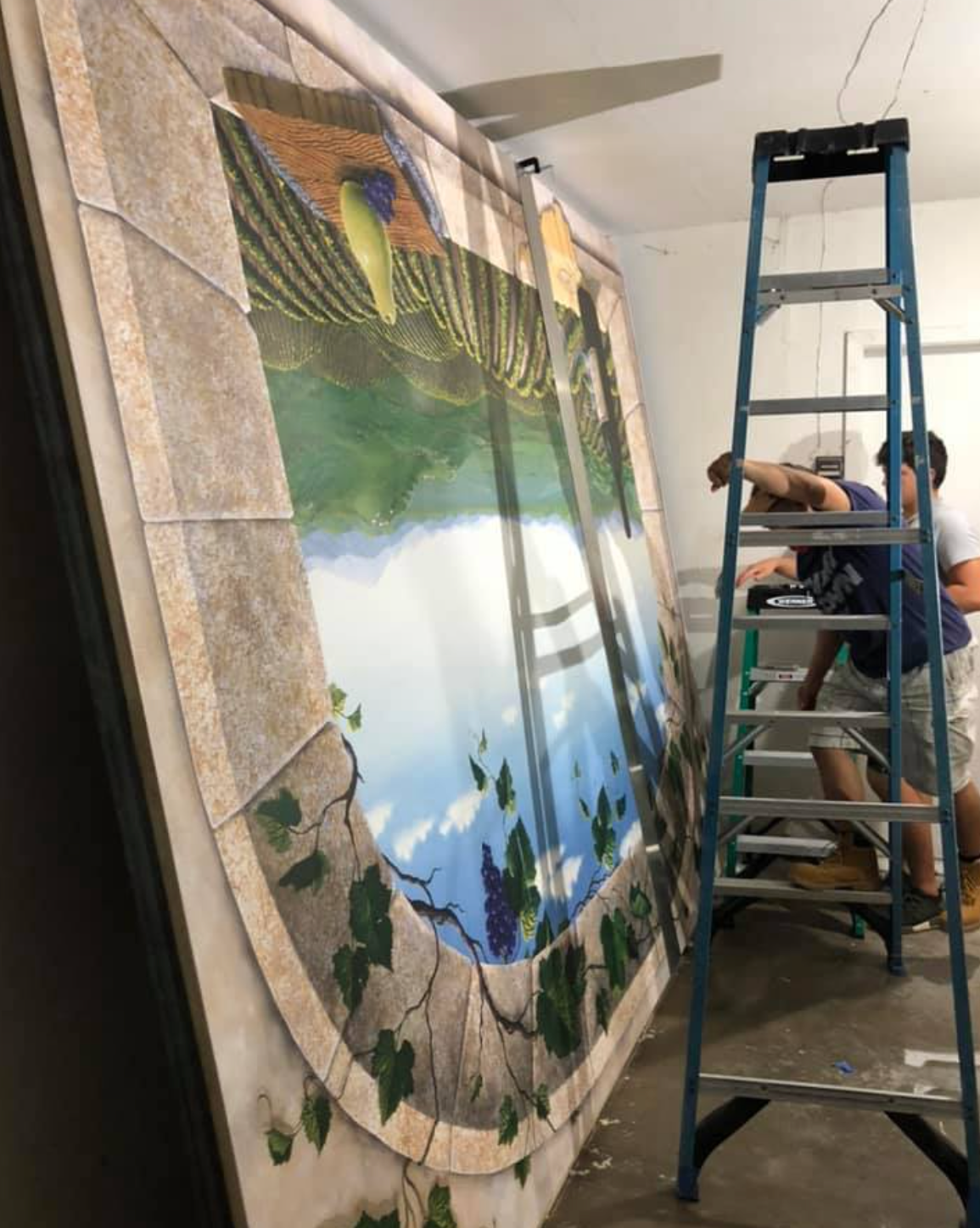 Two people install a large painted mural of a vineyard scene. One on a ladder, working on it.