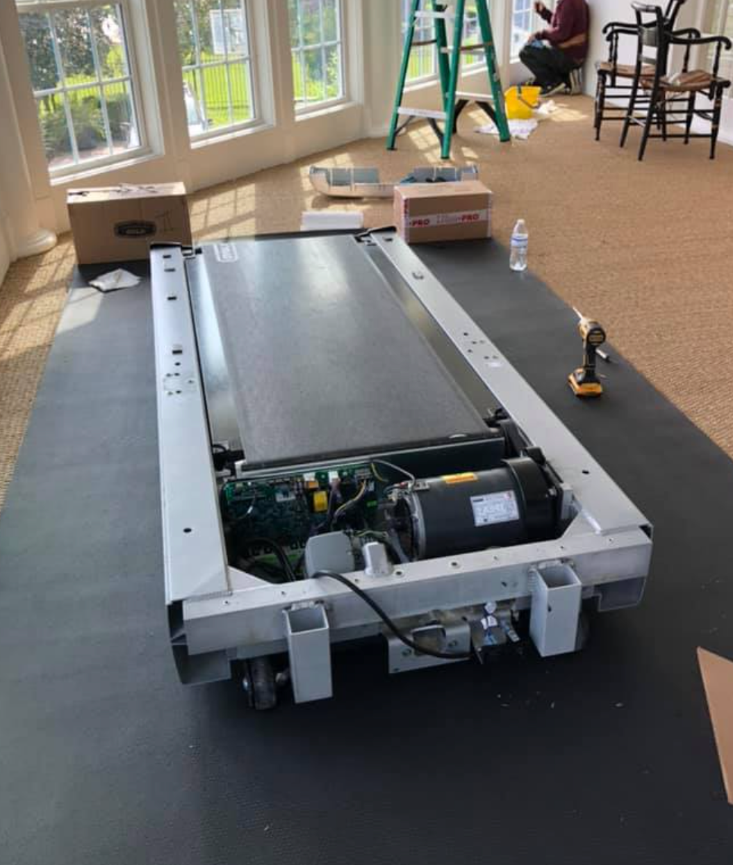 Treadmill disassembled on black mat in a room with windows; person working in the background.