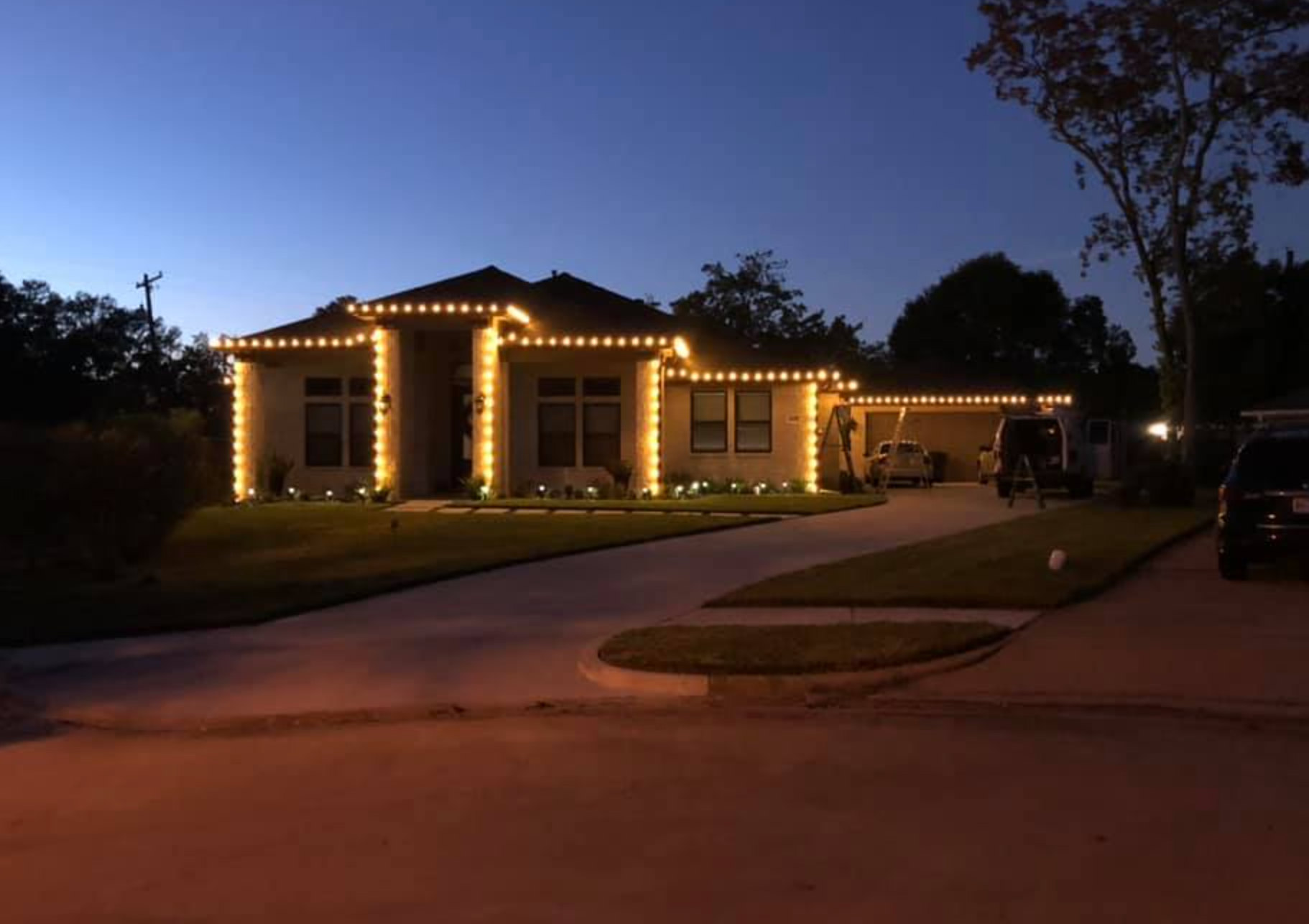 House at dusk with warm white Christmas lights outlining the roof, windows, and bushes.