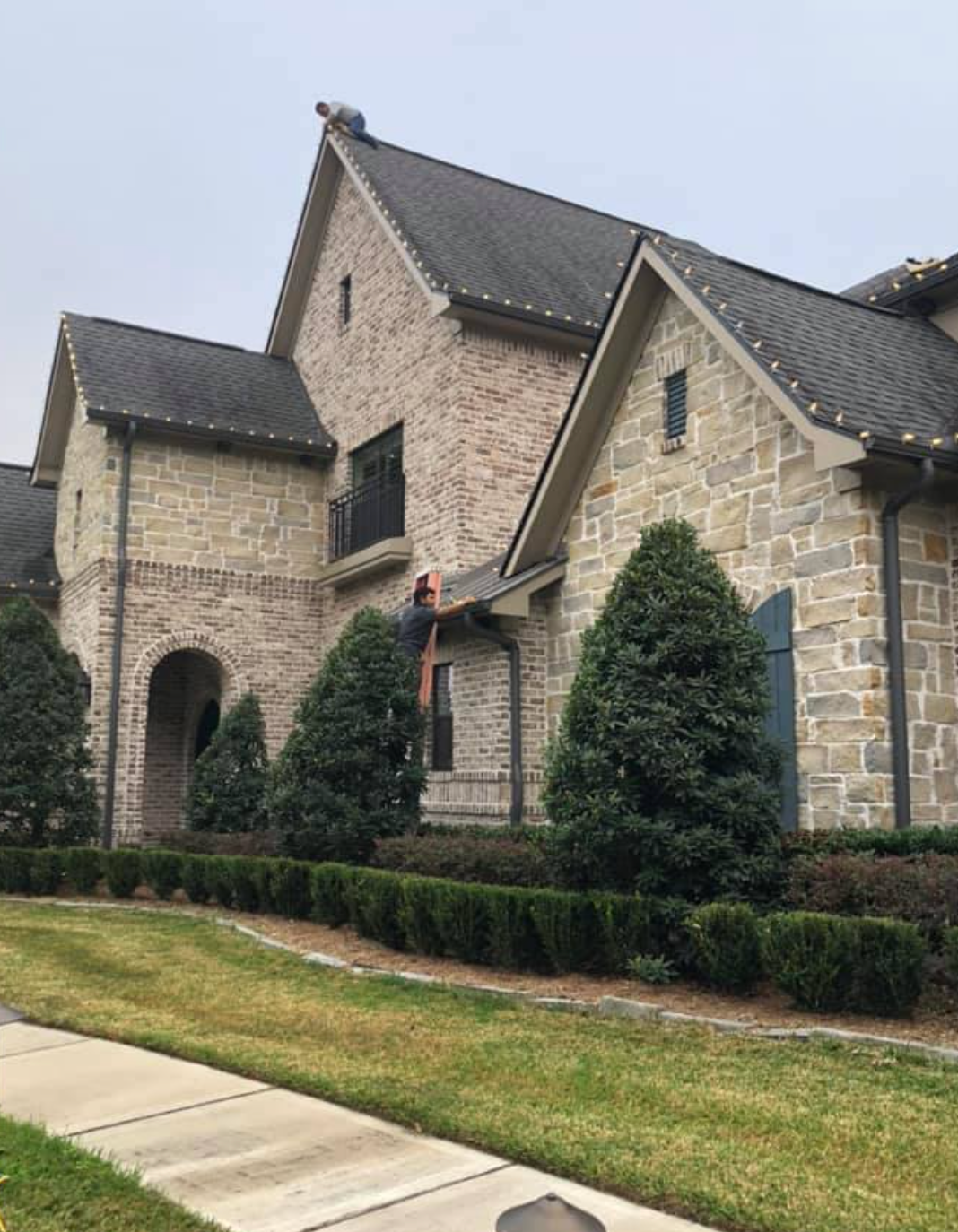 Person on a ladder cleaning gutters of a large, brick house. Green bushes and lawn in front. Overcast sky.