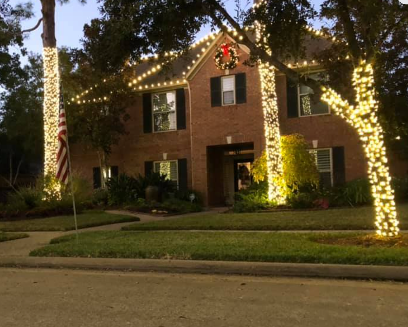 A two-story brick house decorated with Christmas lights on trees and roof. A wreath hangs above the door.