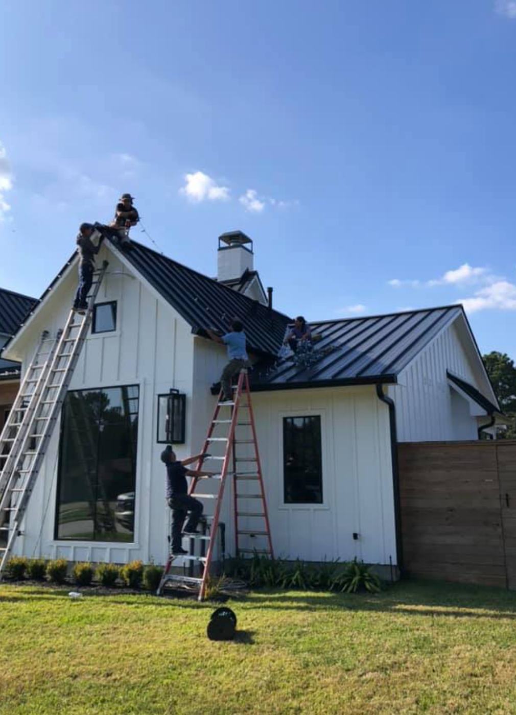 Workers on a white house roof installing black metal panels on a sunny day. Ladders are used for access.
