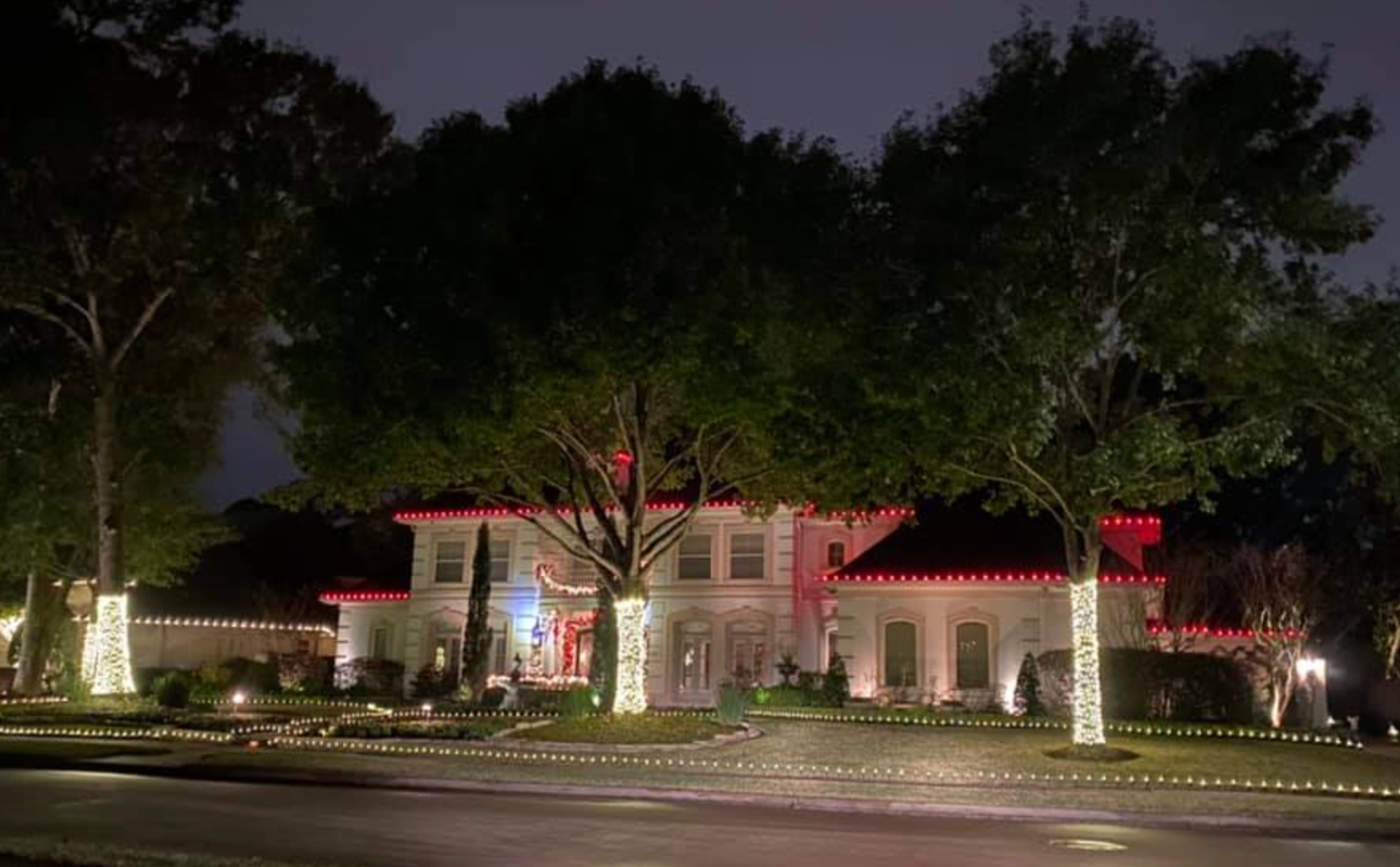 A large, white house at night, decorated with red and white Christmas lights. Trees are also decorated.