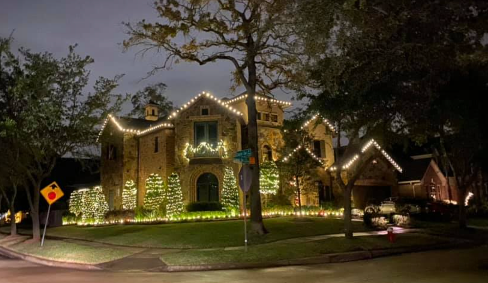 House decorated with white Christmas lights. Exterior view at dusk. Trees and stop sign in the foreground.