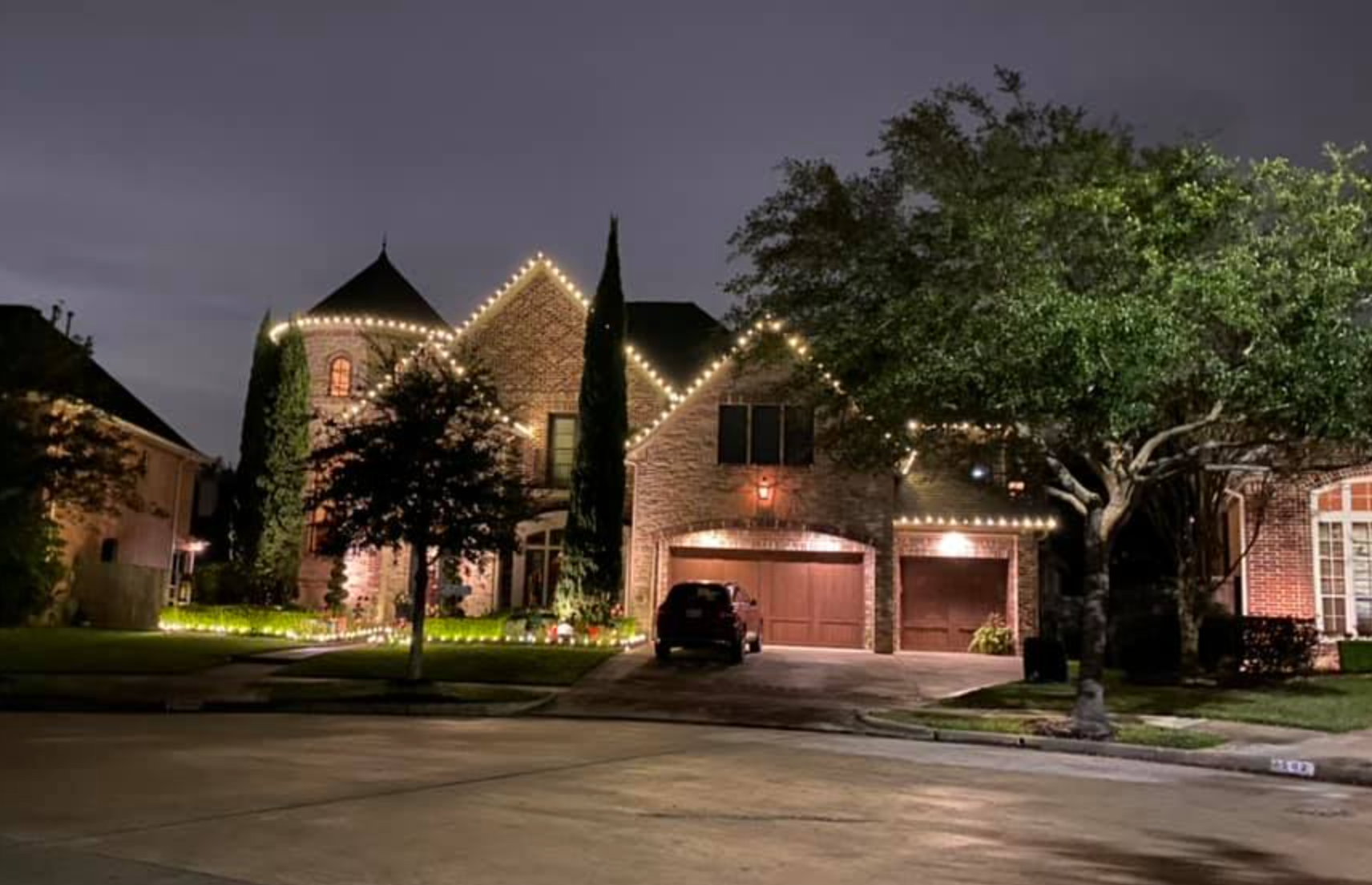 House at night, illuminated with white lights. Garage doors closed, car in driveway, trees to the side.