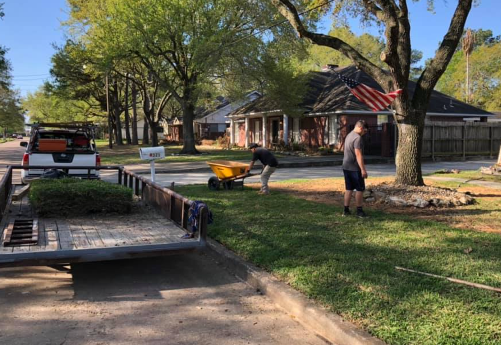 Two men spreading fertilizer on a lawn. Trailer and truck parked on the side of the road in a residential neighborhood.