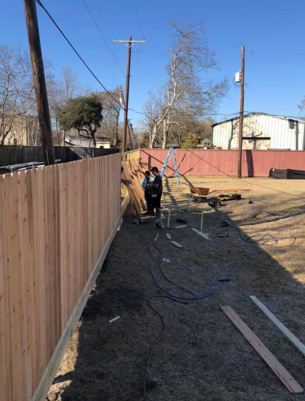 Workers building a wooden fence along a property line, with utility poles overhead.