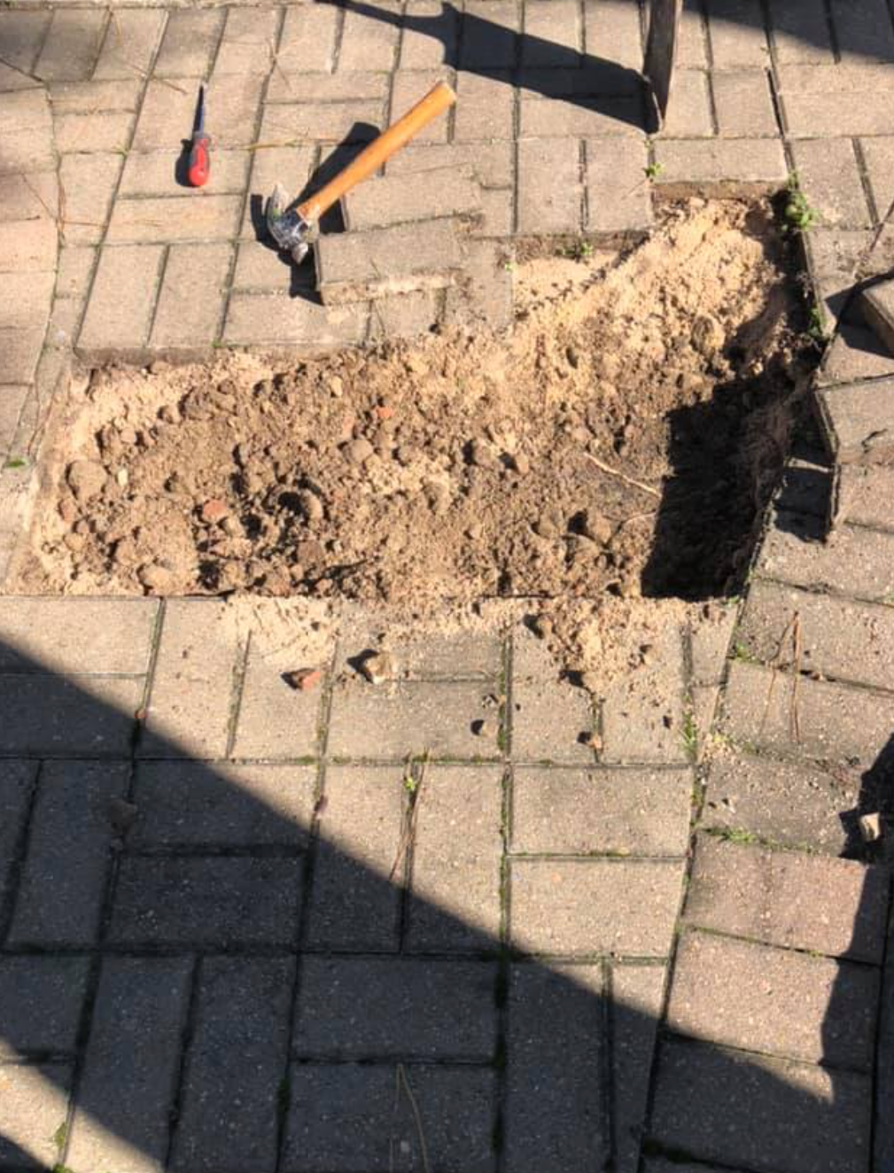 A rectangular section of paving stones is removed, revealing sand, with a hammer and tool nearby.