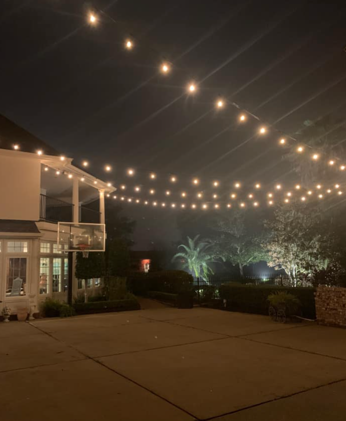 String lights illuminate a backyard at night, spanning between a house and trees.