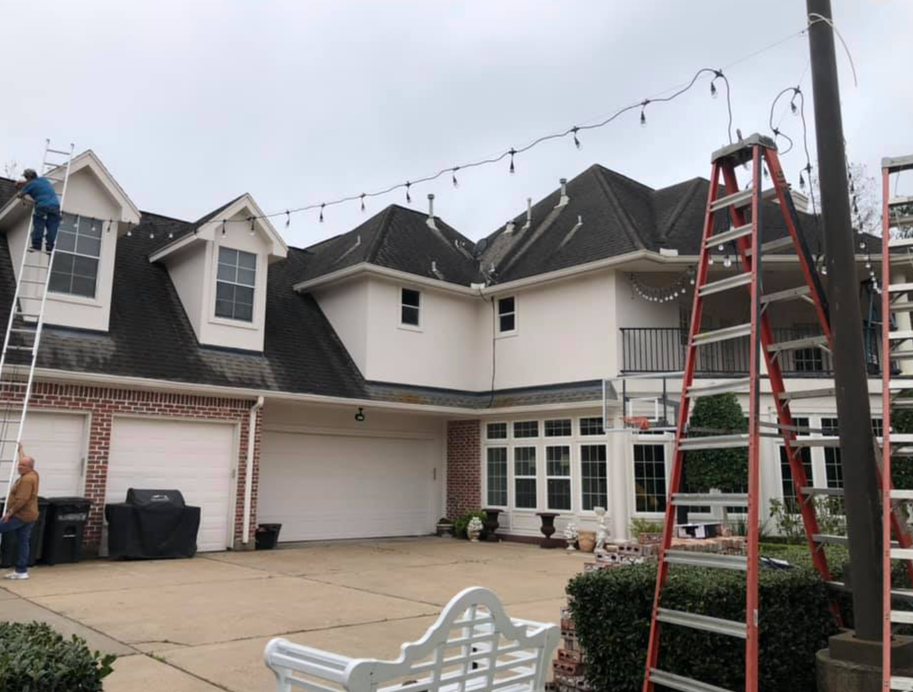 People hanging string lights on a two-story house with a dark roof, using ladders. Overcast sky.